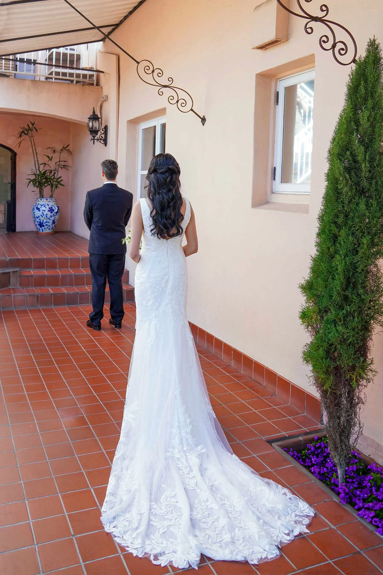 A photograph capturing a "first look" moment between a bride and groom in an outdoor courtyard. The bride stands in the foreground, viewed from behind, wearing a white lace wedding dress with a long train. The groom stands a short distance away on a 