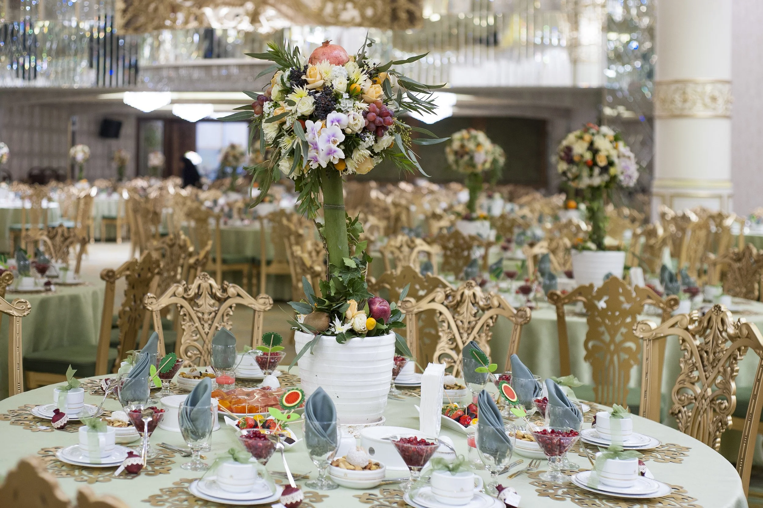 An elegant, formal banquet hall set up for an event, featuring round tables with sage green tablecloths, ornate gold chiavari chairs, and elaborate floral and fruit centerpieces under warm lighting.
