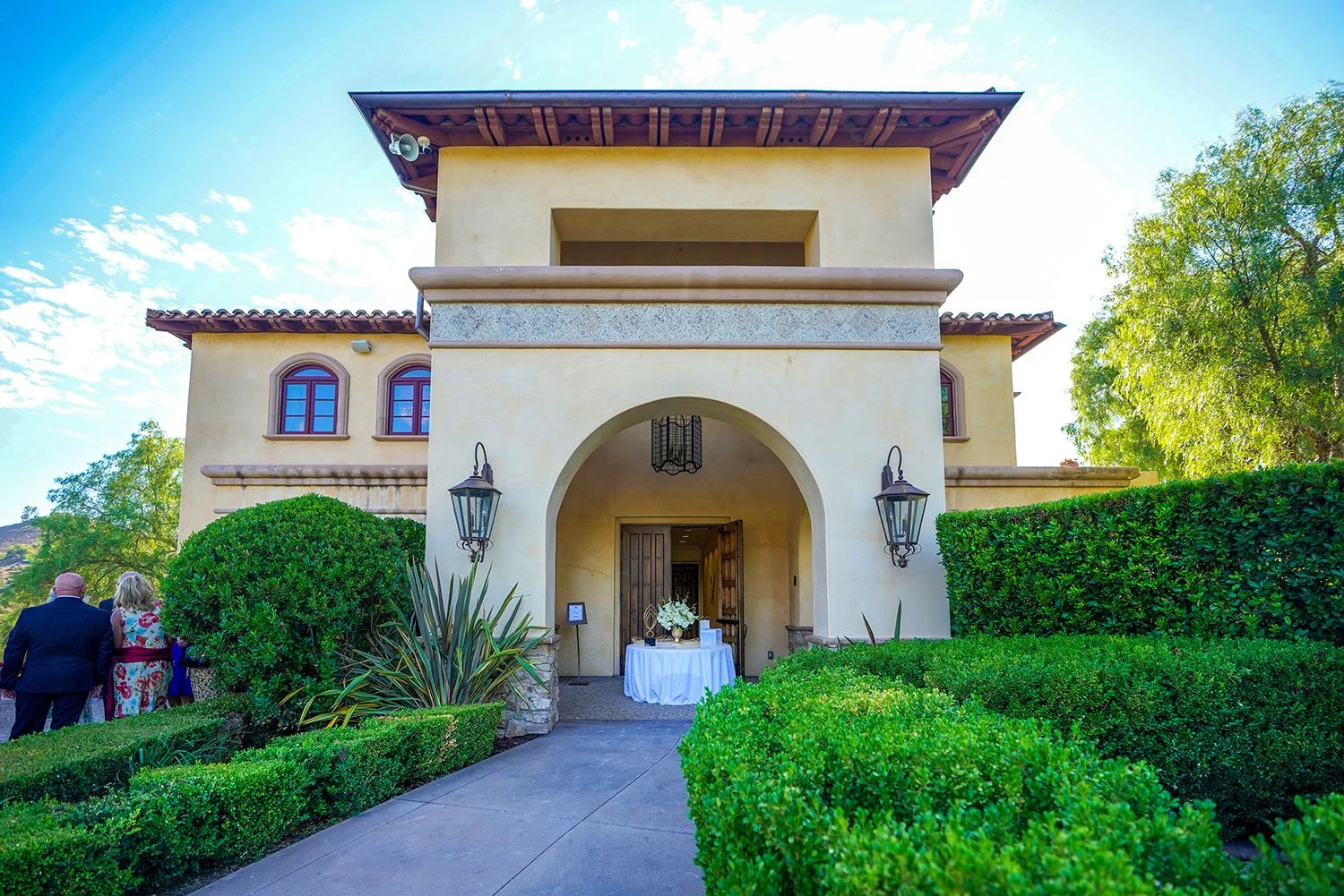 The Mediterranean-style entrance to a building, possibly the Maderas Golf Club clubhouse, featuring an arched entryway with a wooden door, a small table covered in a white cloth, a path lined with manicured green hedges, large dark lanterns, a couple