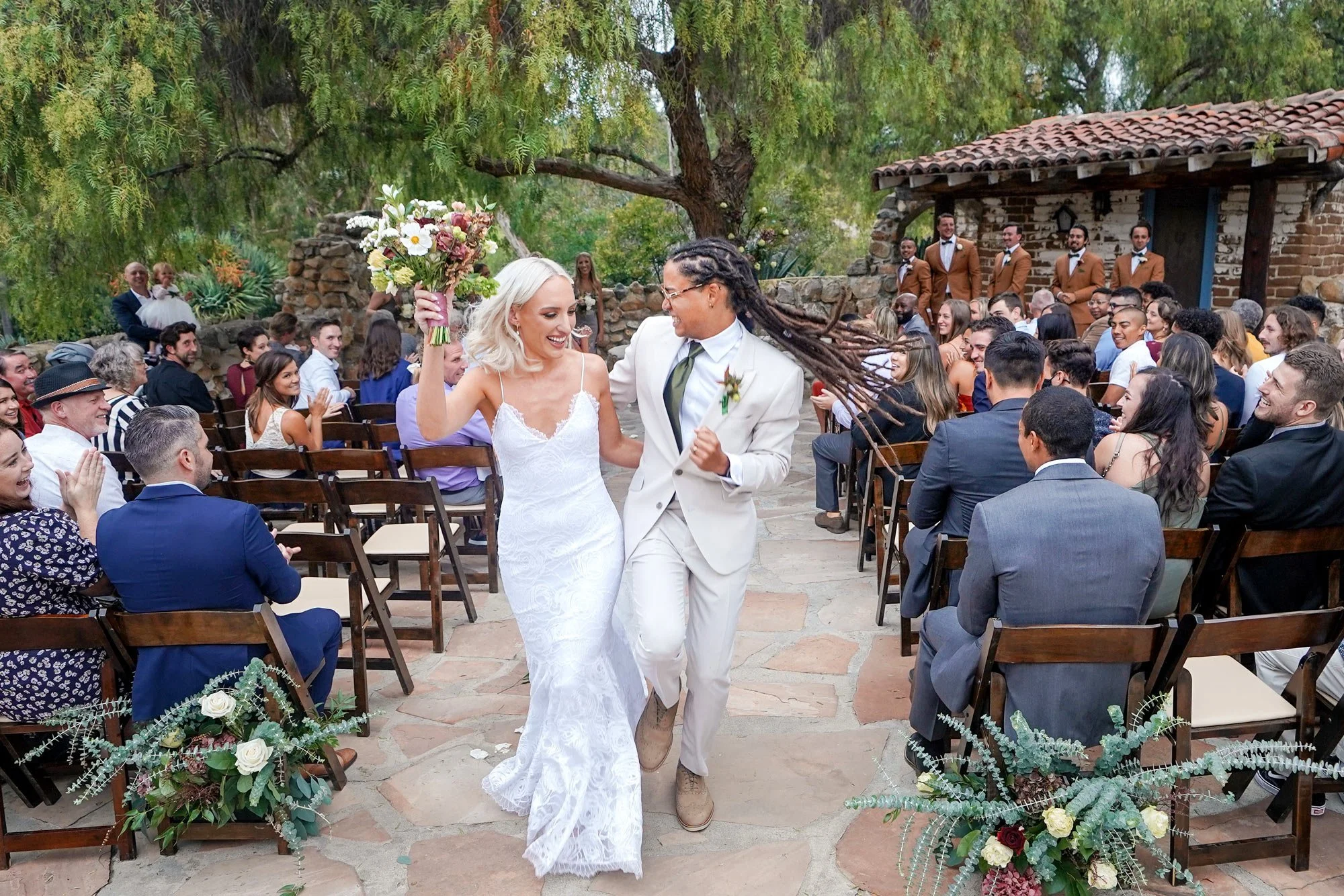 A photo of a newly married couple walking cheerfully down an outdoor aisle after their wedding ceremony, with the bride in a white lace dress holding a bouquet and the groom in a cream-colored suit, as seated guests cheer them on in Leo Garrillo Ranc