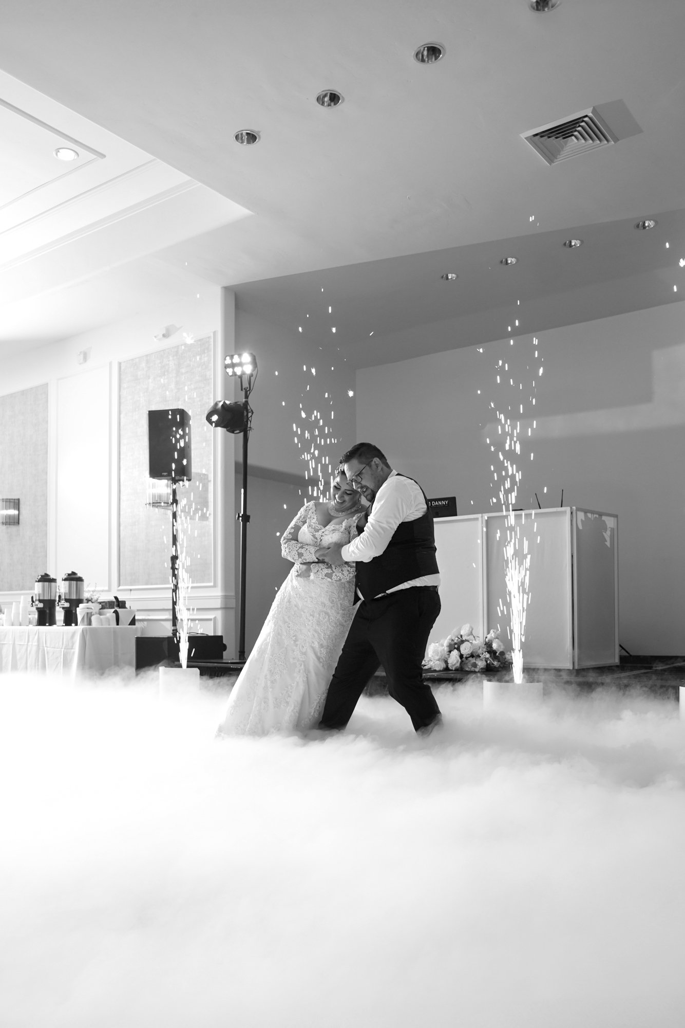 A black and white photograph capturing a wedding reception first dance. The bride in a white lace dress and the groom in a dark vest over a white shirt embrace and dance on a floor covered in low-hanging white fog, with indoor sparks or fireworks goi