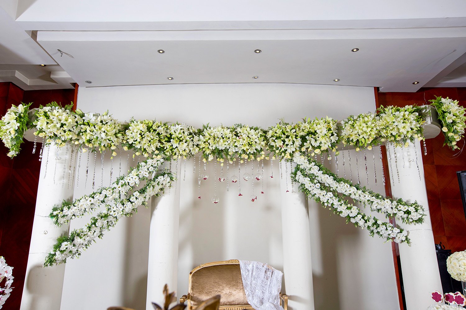 An elaborate indoor event display featuring a large horizontal swag of white flowers and hanging crystal accents, with additional white flower arrangements draped down to form an arch around a gold chair in a wall niche.