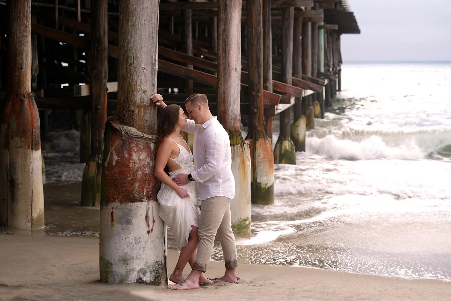 An outdoor engagement session photograph of a couple embracing and about to kiss on a sandy beach in Pacific Beach, standing under a large wooden pier as waves crash nearby. The woman wears a white sundress, and the man wears a white shirt and tan pa