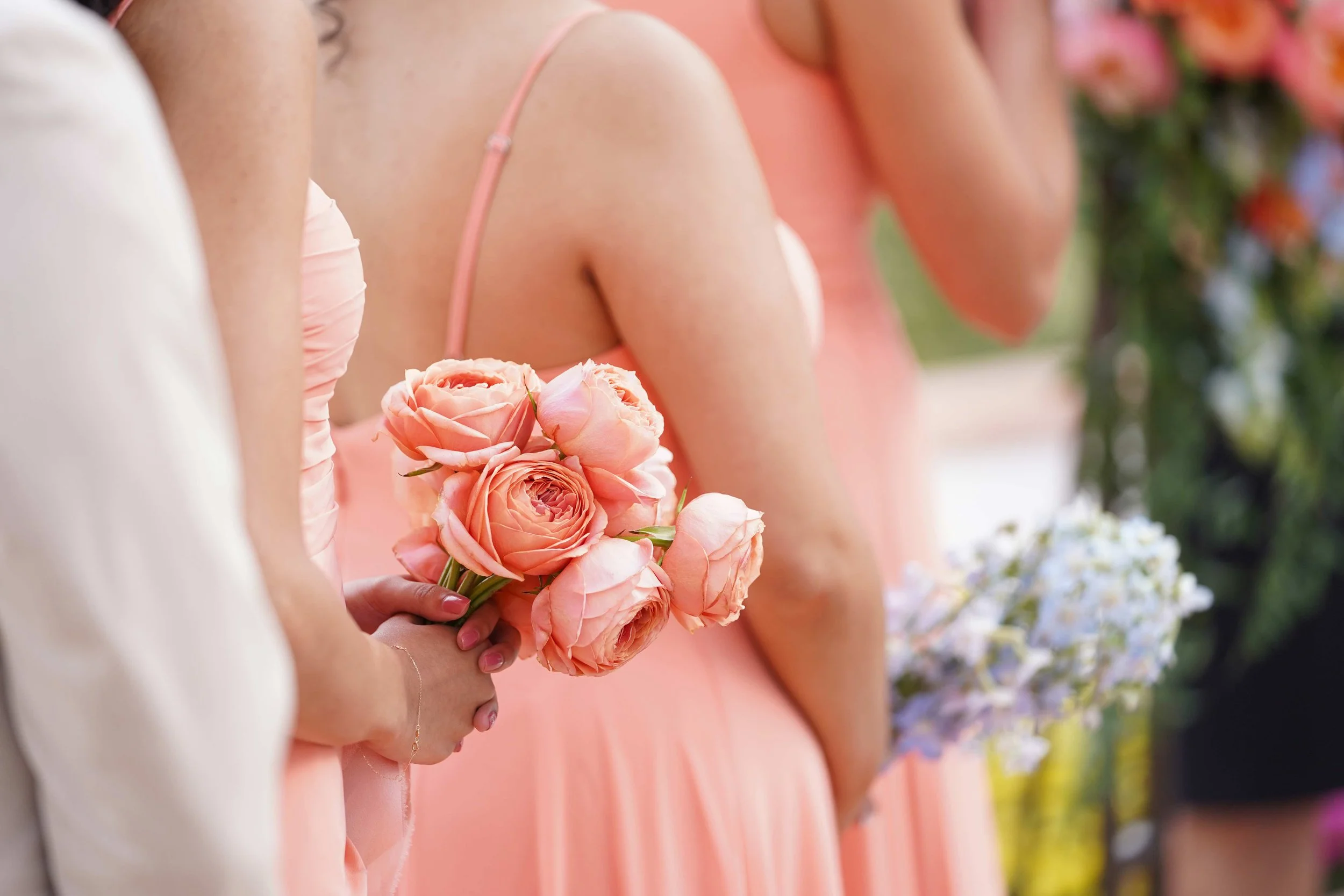A close-up shot of a bridesmaid's hand holding a small bouquet of peach-colored roses, standing beside another bridesmaid in a matching dress with a bouquet of light blue flowers nearby.



