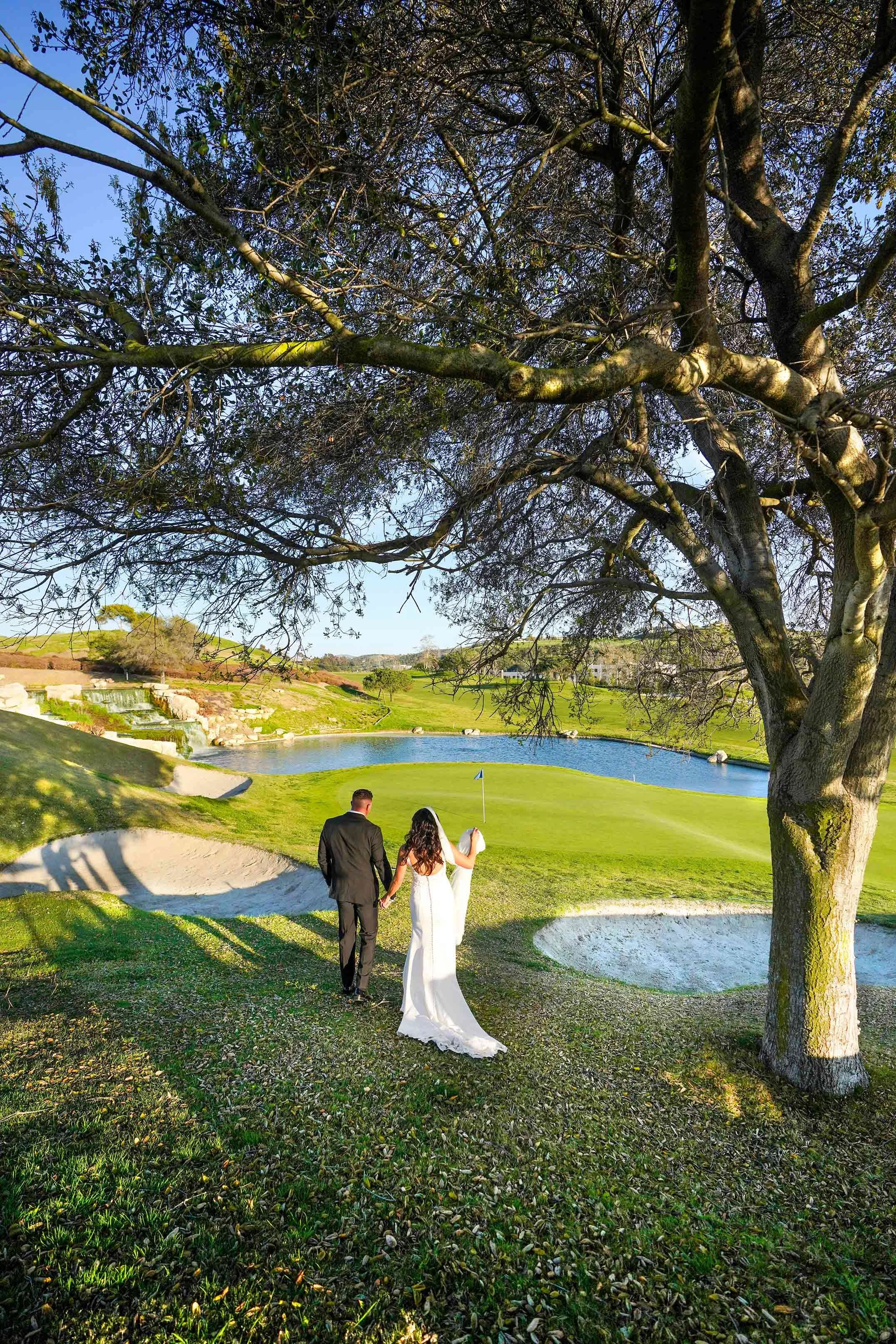 An outdoor photograph of a newly married couple walking hand-in-hand down a grassy slope on a sunny day. The groom wears a black suit and the bride wears a white wedding dress with a long train. They are viewed from behind, walking past sand bunkers 