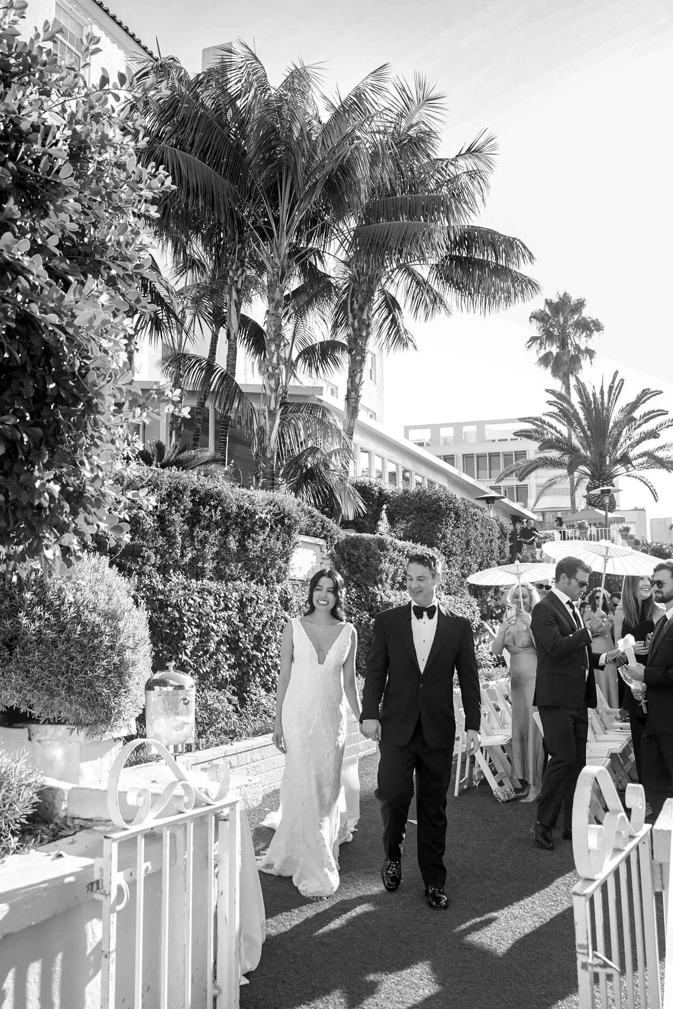 A black and white outdoor photograph of a newly married couple walking down an aisle after their wedding ceremony. The bride wears a white sleeveless lace wedding dress and holds a white floral bouquet. The groom wears a black tuxedo with a black bow