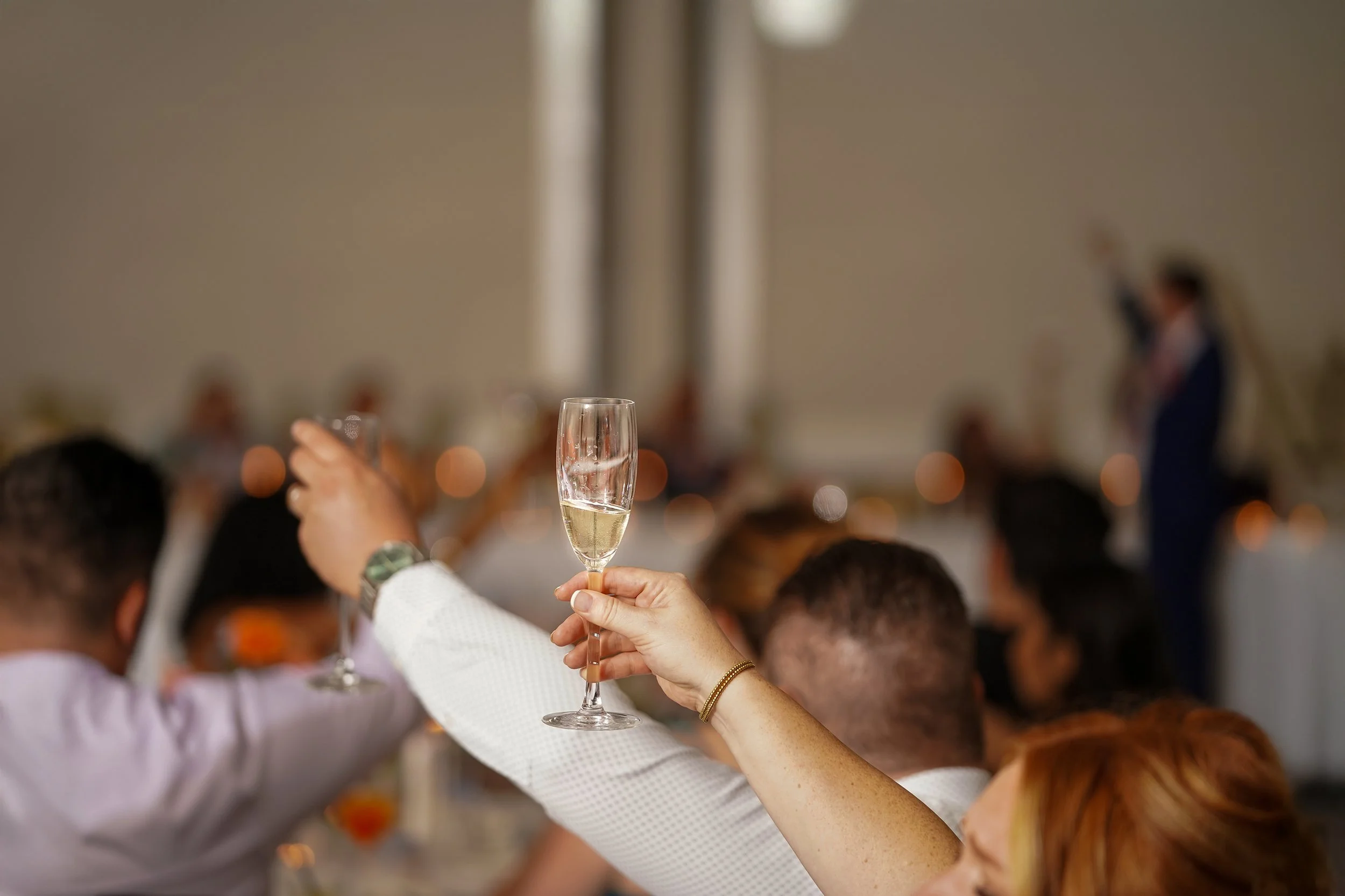 A close-up, shallow-focus shot of a person’s hand raising a tall champagne flute during a wedding toast. In the blurred background, other guests also raise their glasses in celebration at a long, white-clothed reception table.