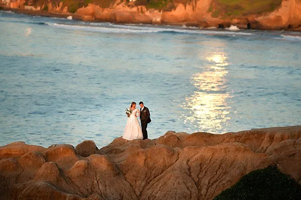 A bride and groom stand together on a large, textured rocky cliffside overlooking a vast, shimmering blue ocean. The sunlight reflects brightly off the water's surface as waves crash against the shoreline and distant cliffs in the background.