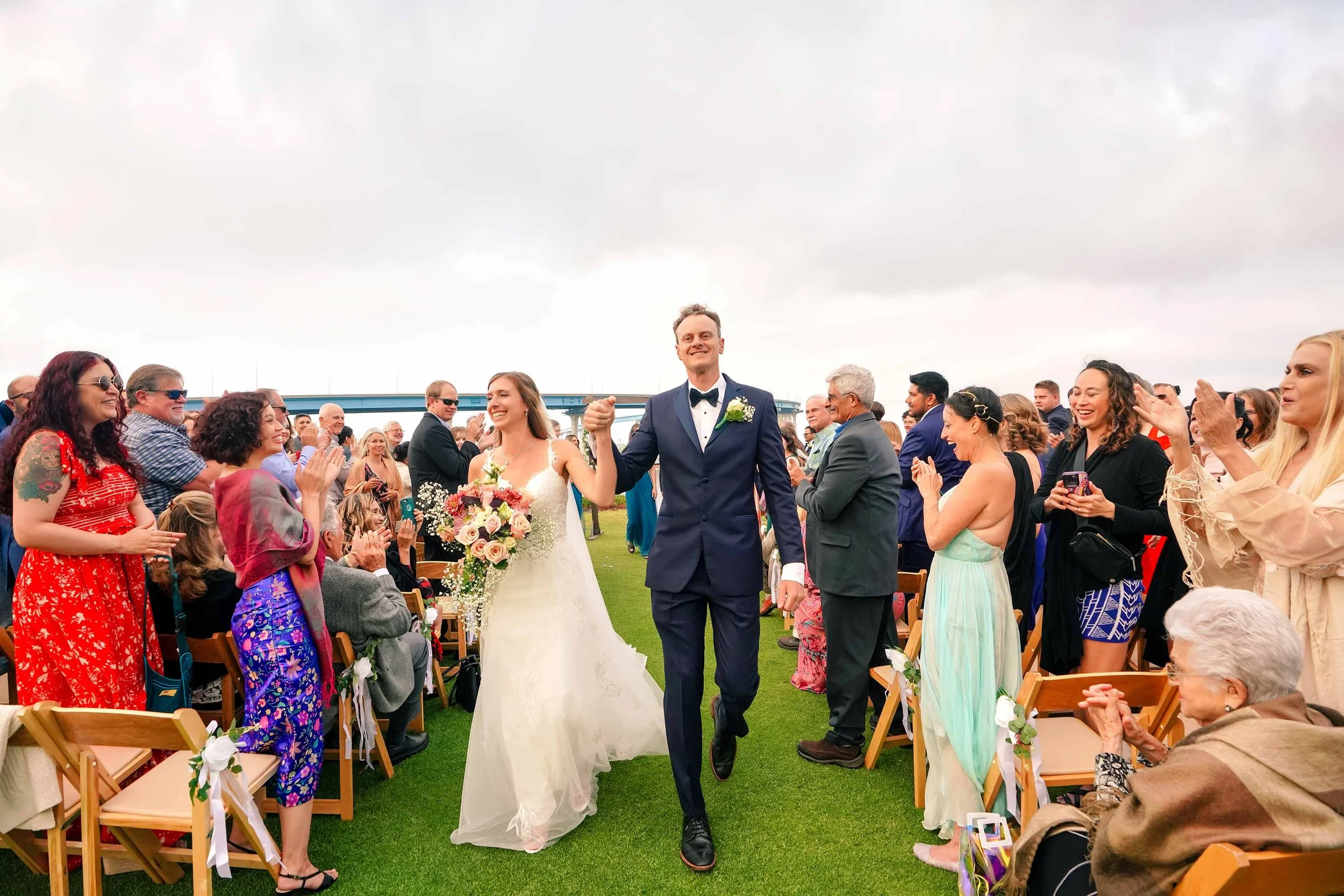 A joyful bride in a white lace gown holding a pink and white floral bouquet and a groom in a navy blue tuxedo and bow tie walk down an outdoor wedding aisle as guests in white folding chairs smile and applaud in San Diego.


