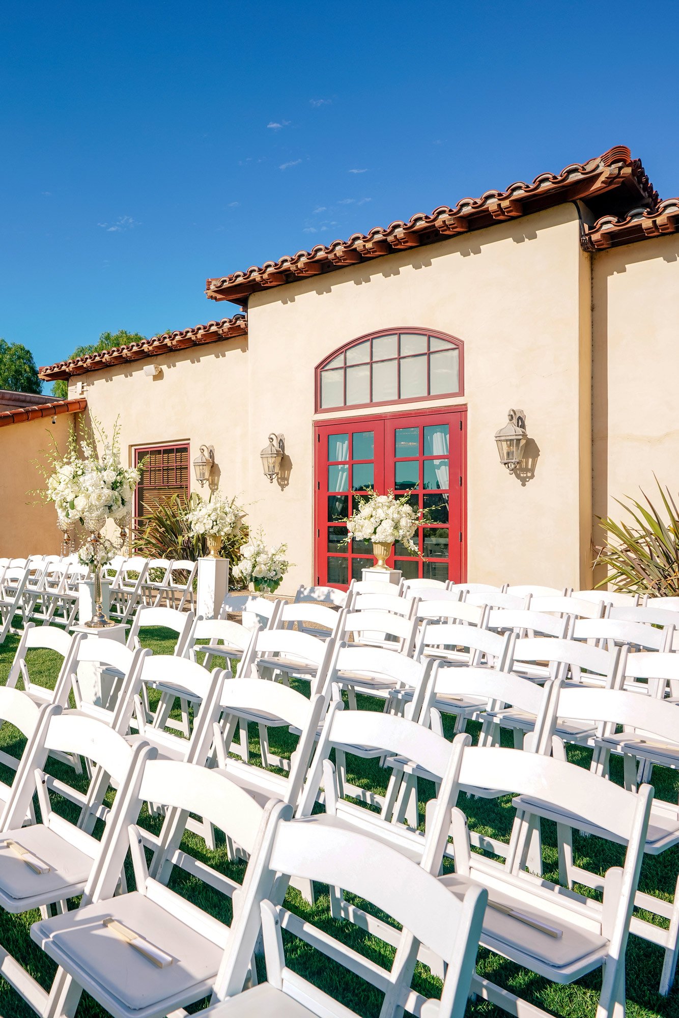 A bright outdoor photograph of an empty wedding ceremony setup on a grassy lawn. Numerous rows of white folding chairs face a Spanish-style stucco building with a red-tiled roof and vibrant red double doors at Maderas Golf Club. Large white floral ar