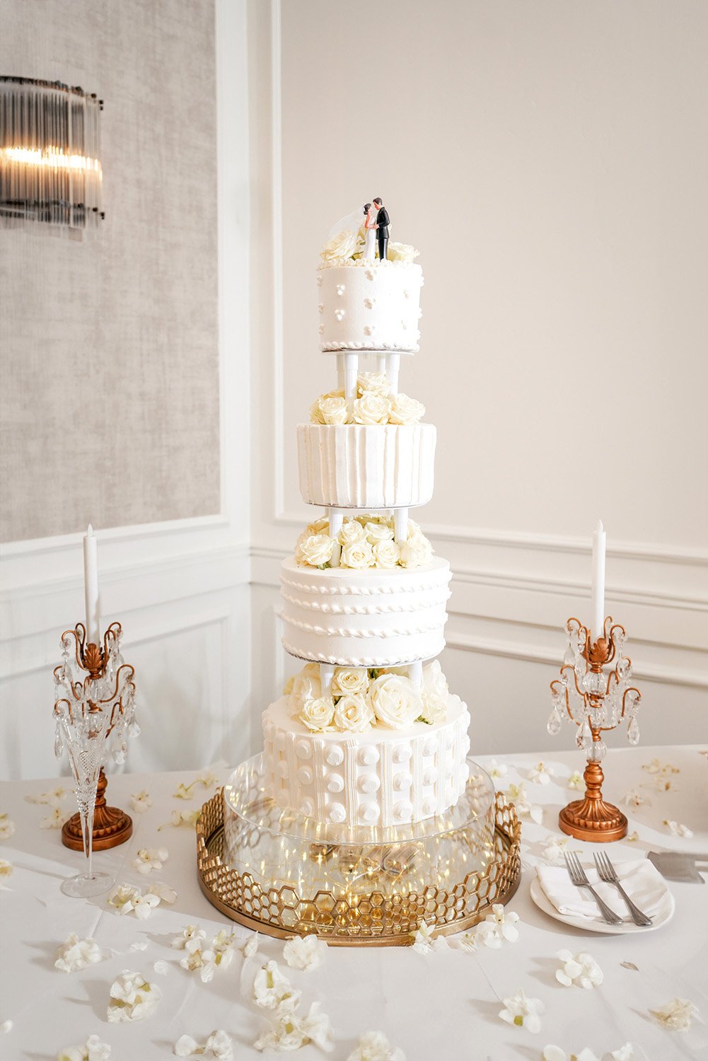 A tall, four-tiered white wedding cake adorned with white roses and a bride and groom topper. The cake sits on a clear stand, placed on a gold decorative serving tray on a white-clothed table, scattered with white flower petals. Two crystal and coppe