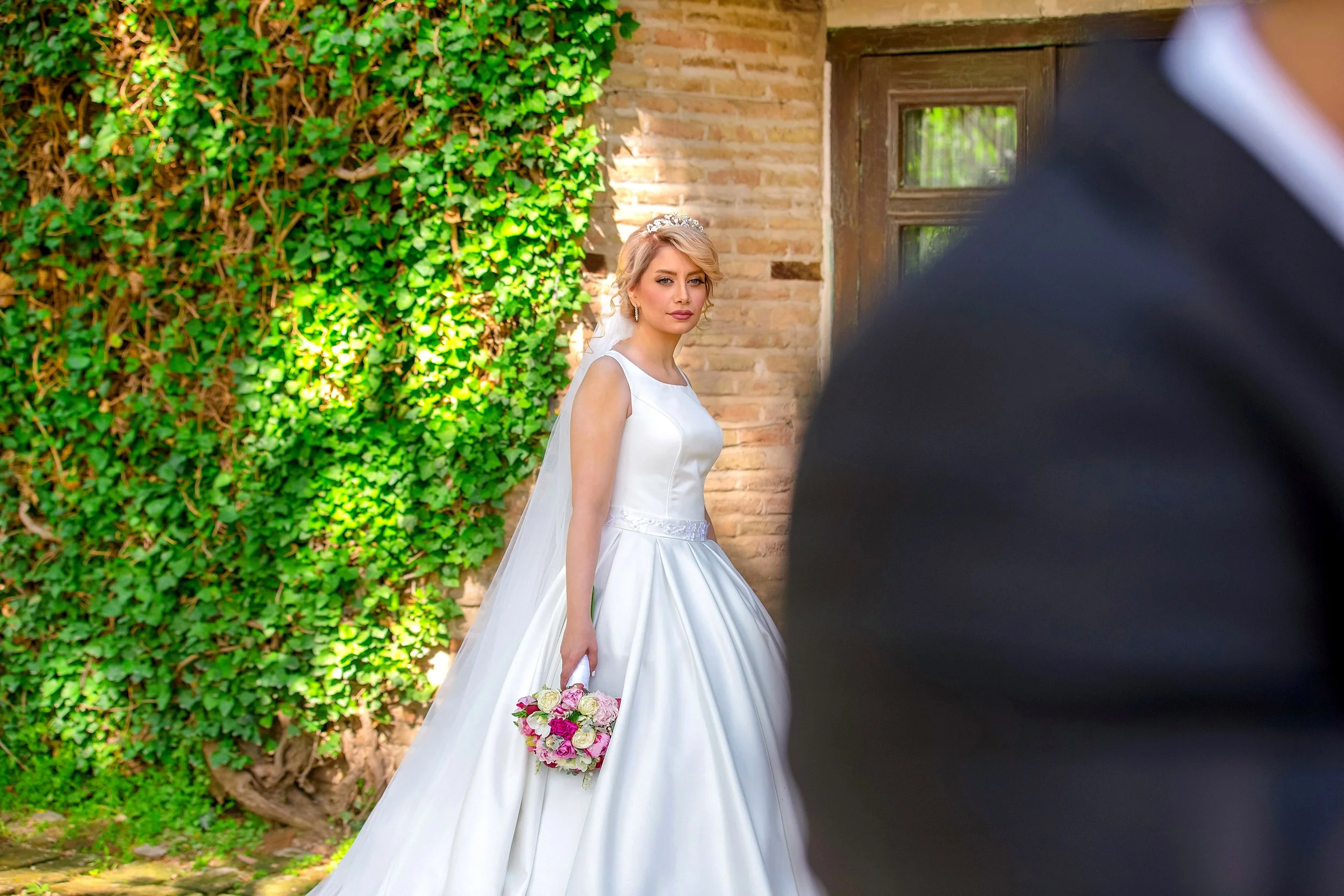 A bride in a white wedding dress holding a bouquet looks towards the camera while the groom stands nearby in a suit.
