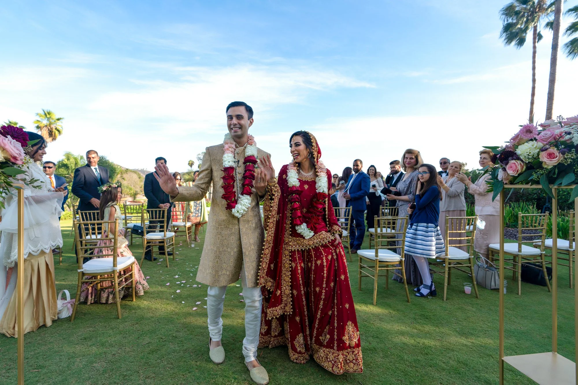 An outdoor photograph of a newly married bride and groom smiling as they walk down the aisle after their wedding ceremony. The groom wears a gold sherwani and the bride wears an ornate, deep red lehenga with gold embroidery, both adorned with red and
