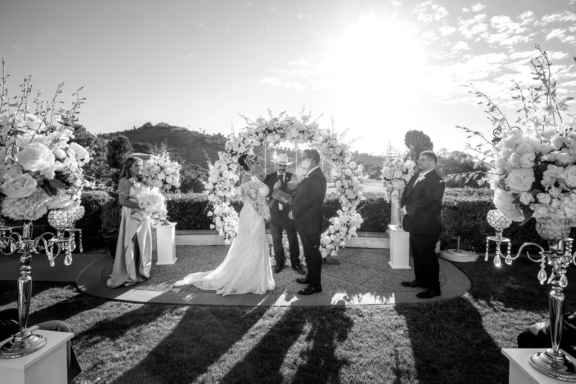 A black and white outdoor photograph of a wedding ceremony. The bride in a white lace dress and the groom in a dark tuxedo stand at the altar under a white floral arch. An officiant is between them, and a woman in a light dress and a man in a dark su