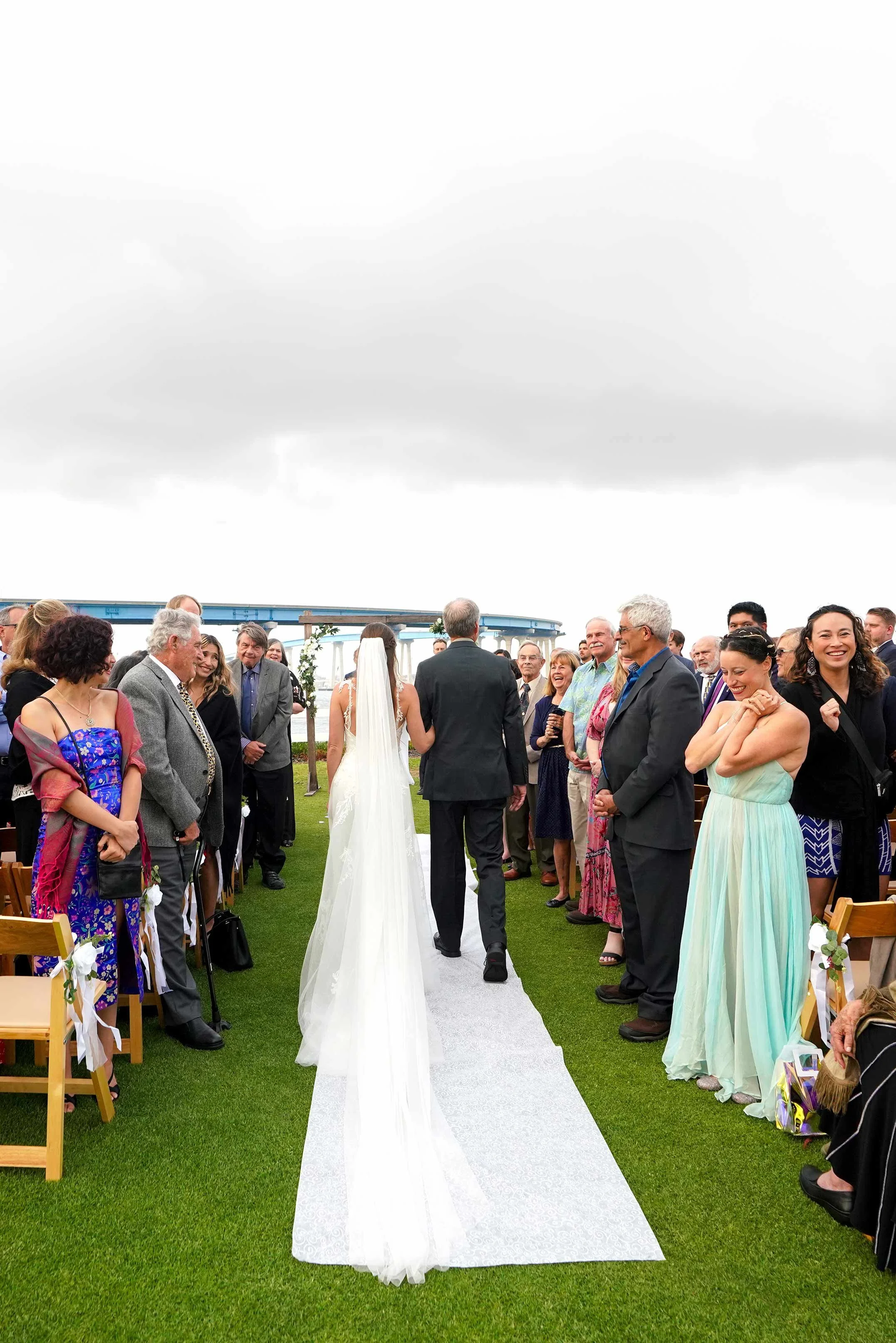 A view from behind a bride in a white gown and her father in a dark suit walking together down a white aisle runner on a green lawn during an outdoor wedding ceremony in Coronado, with seated guests watching on both sides.


