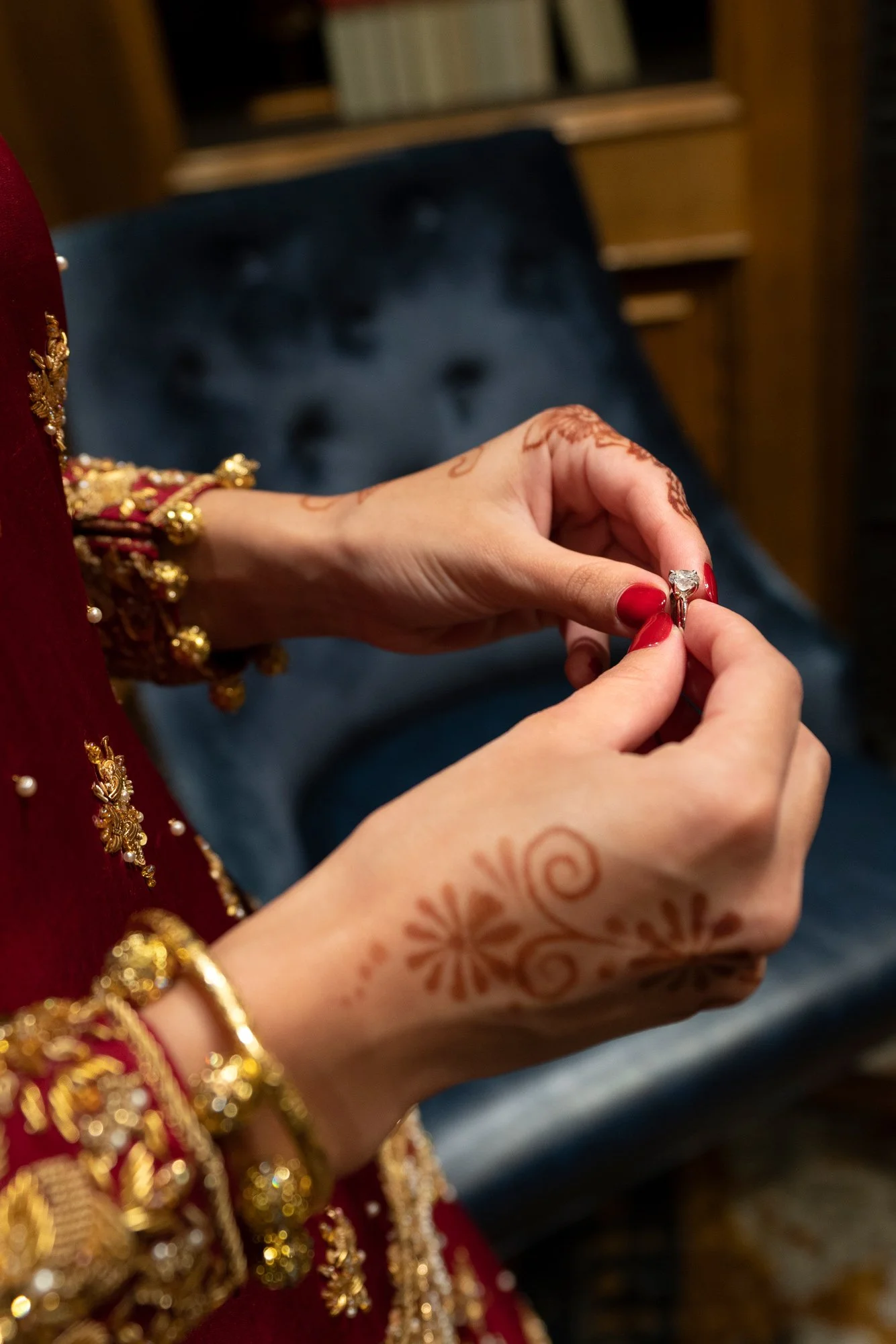A close-up photograph of a person's hands, adorned with henna tattoos and red nail polish, holding a diamond ring. The person is wearing a deep red outfit with gold embroidery and a gold bangle on their wrist. A blurred, dark blue velvet chair is in 