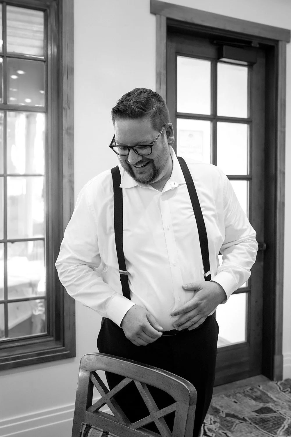 A groom indoors, smiling as he adjusts his white shirt and black trousers while wearing black suspenders and glasses. He stands in a room with a wooden chair, a glass door, and a window.