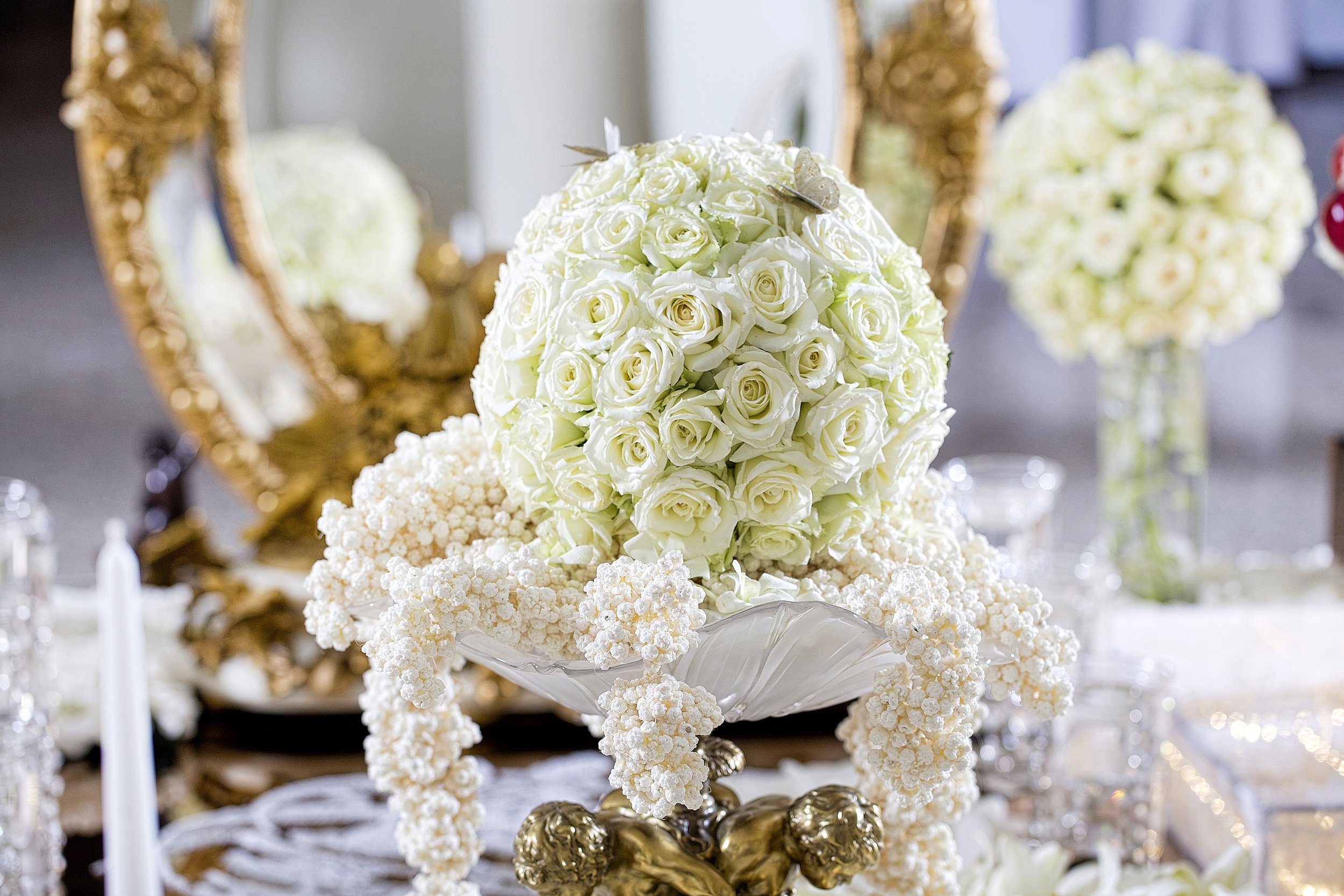 An ornate floral centerpiece featuring a sphere of white roses and draped white flower garlands, displayed on a gold stand with a large gold mirror and another floral arrangement in the background.
