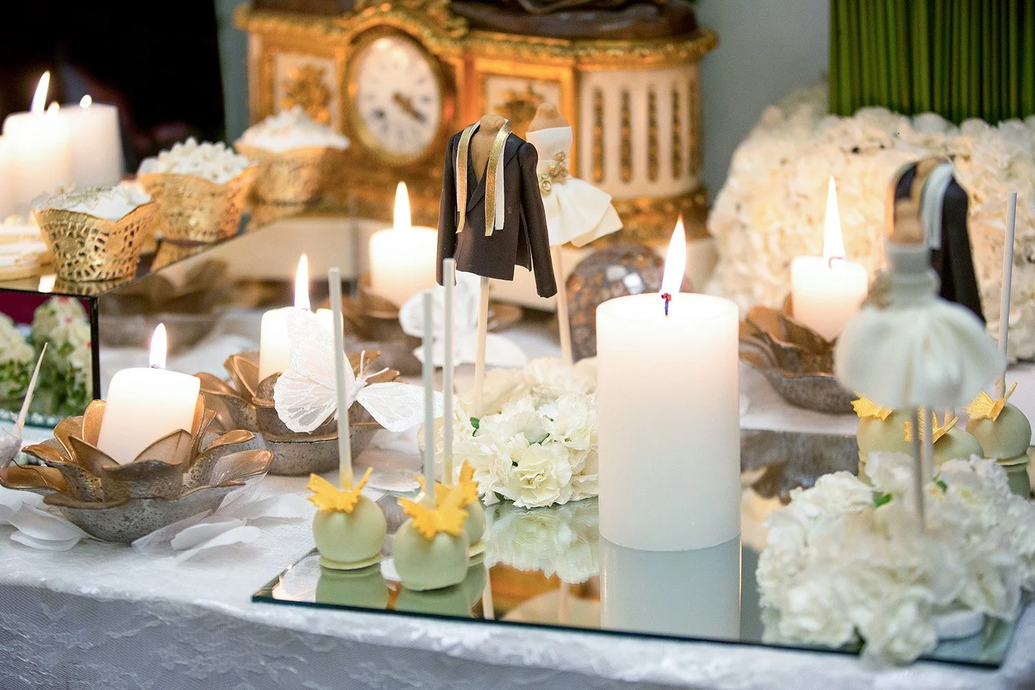The table is draped in white lace or sheer fabric and features a sophisticated color palette of white, gold, and black accents. The soft, warm glow from several pillars and votive candles creates an intimate and romantic atmosphere. The items are dis