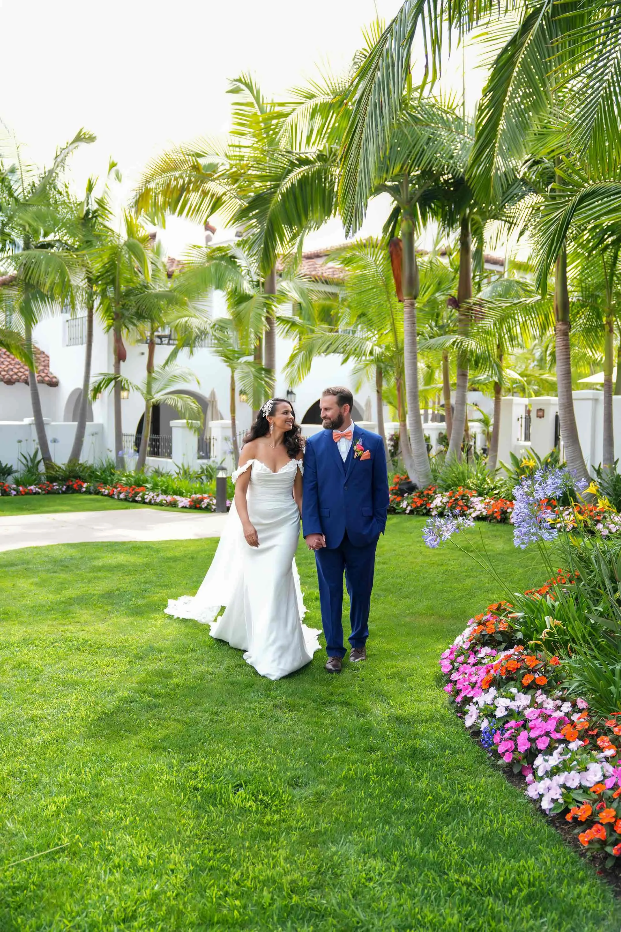 A bride in a white, off-the-shoulder gown and a groom in a bright blue suit with an orange bowtie walk hand-in-hand across a lush green lawn surrounded by tall palm trees and colorful flowers in the Omni Resort, smiling at each other.


