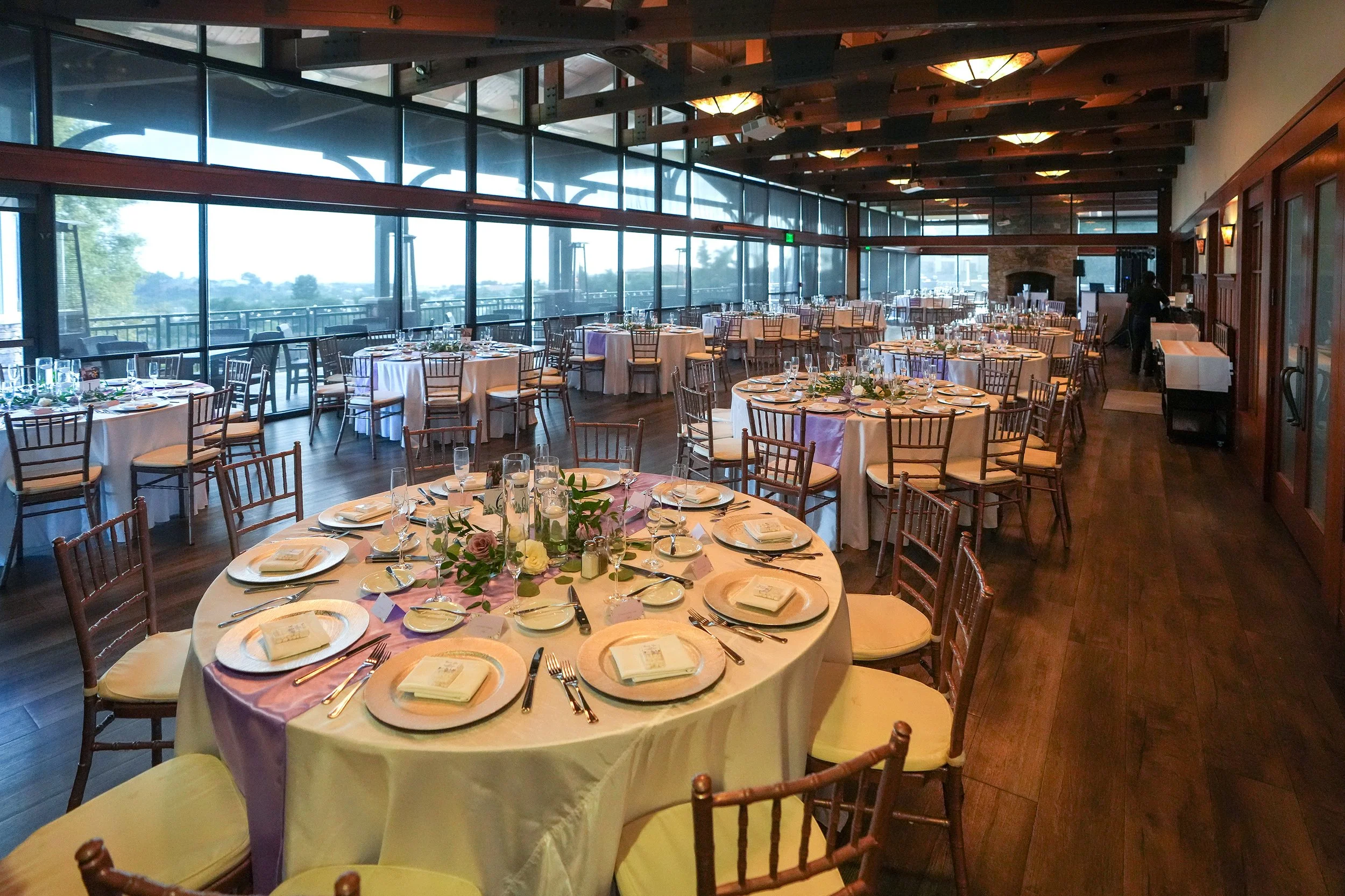 A large, elegant indoor banquet hall or dining room set up for an event, featuring round tables with white and lavender linens, gold charger plates, gold chiavari chairs, exposed wooden beams on the ceiling, and floor-to-ceiling windows with a view o