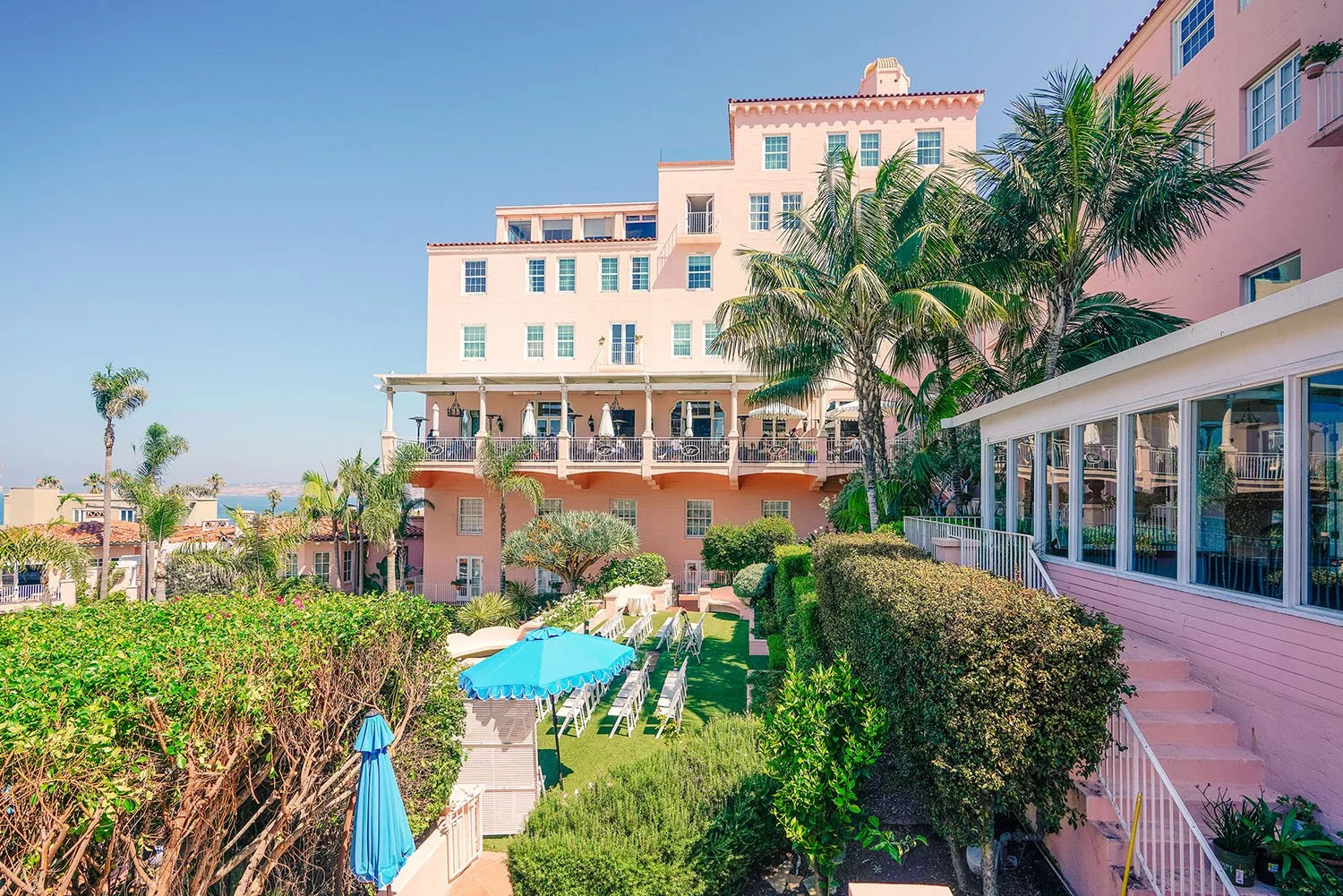 An elevated, daytime view of the multi-story, pink-stucco "Pink Lady" building of the La Valencia Hotel in La Jolla, California, featuring its terraced gardens, palm trees, and ocean views in the distance