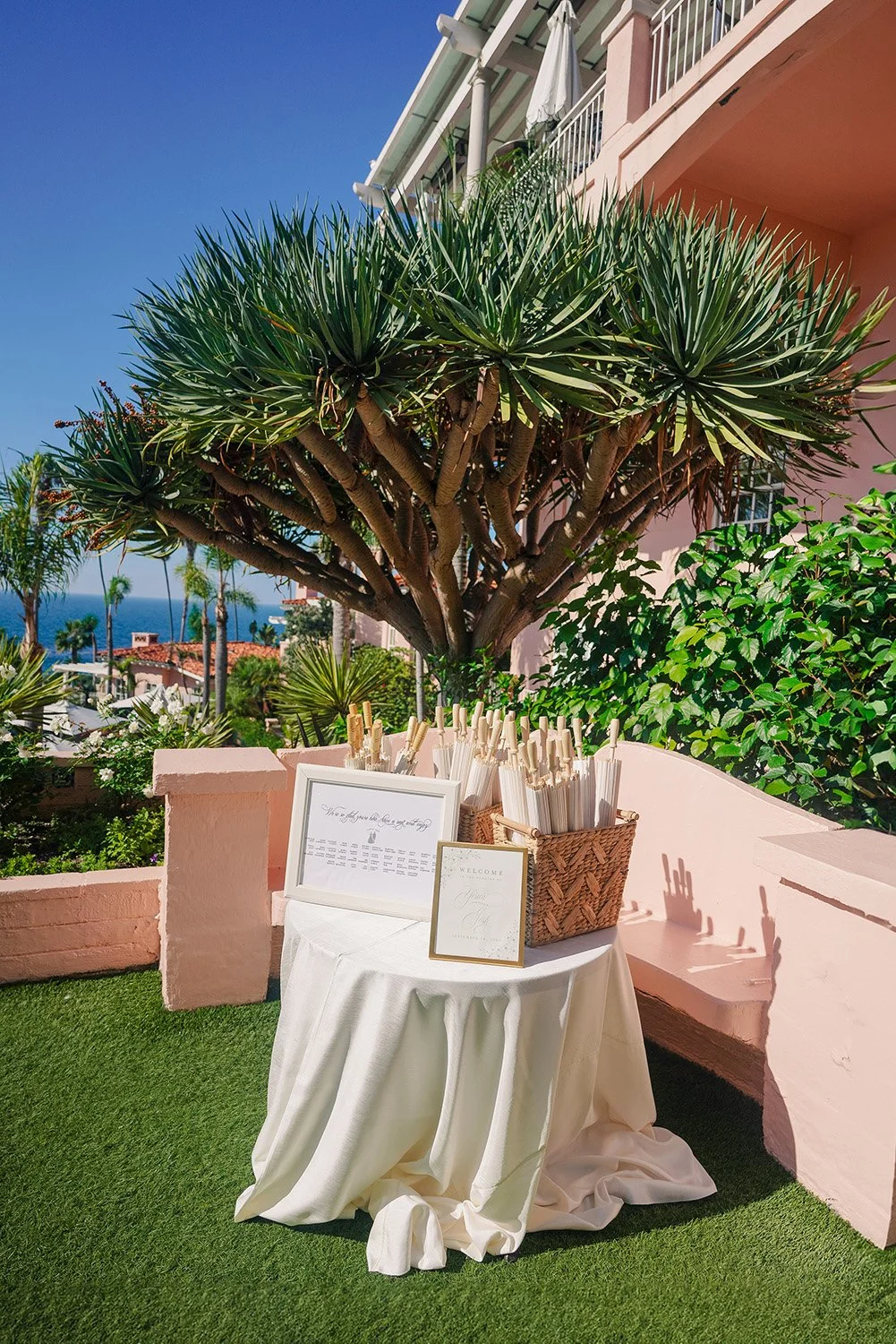 An outdoor event table set up with a white tablecloth, a basket of white favors or fans, and framed signs, all positioned on green turf with a distinctive multi-trunked tree, pink architecture, and an ocean view in the background at the La Valencia H