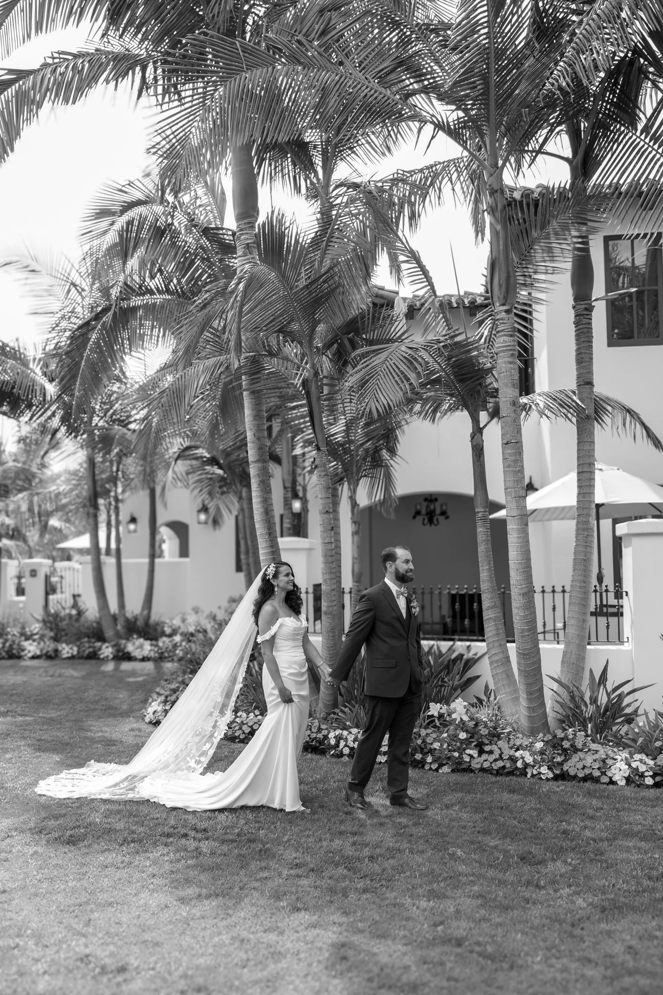 A black and white photograph of a bride in an off-the-shoulder white gown with a long, flowing veil and a groom in a dark suit walking hand-in-hand across a green lawn, surrounded by tall palm trees and white buildings with Spanish-style architecture