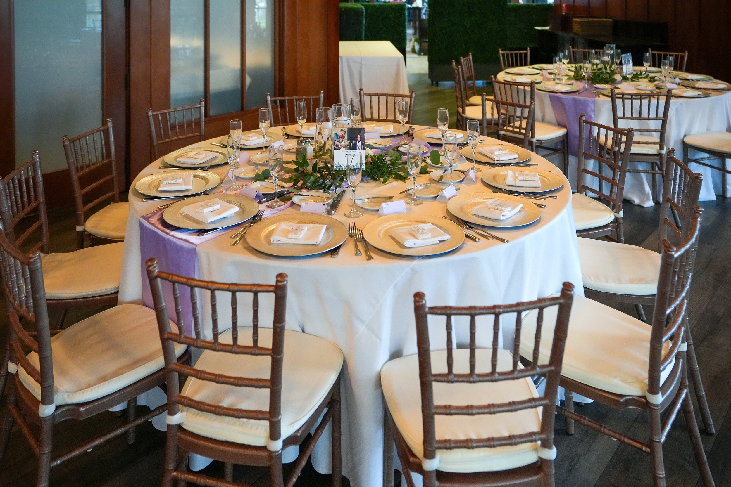 A round table set for a formal dinner event with gold charger plates, lavender fabric accents, white plates and napkins, a green and white floral centerpiece, surrounded by brown wooden chiavari chairs with cream cushions.