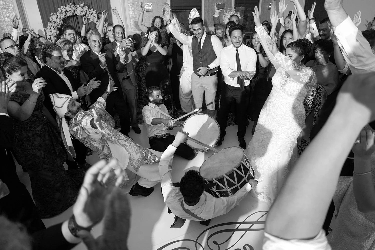 Guests are dancing and cheering around two men playing traditional drums on the dance floor. The bride in a white lace dress and the groom in a dark vest are visible in the celebratory crowd.