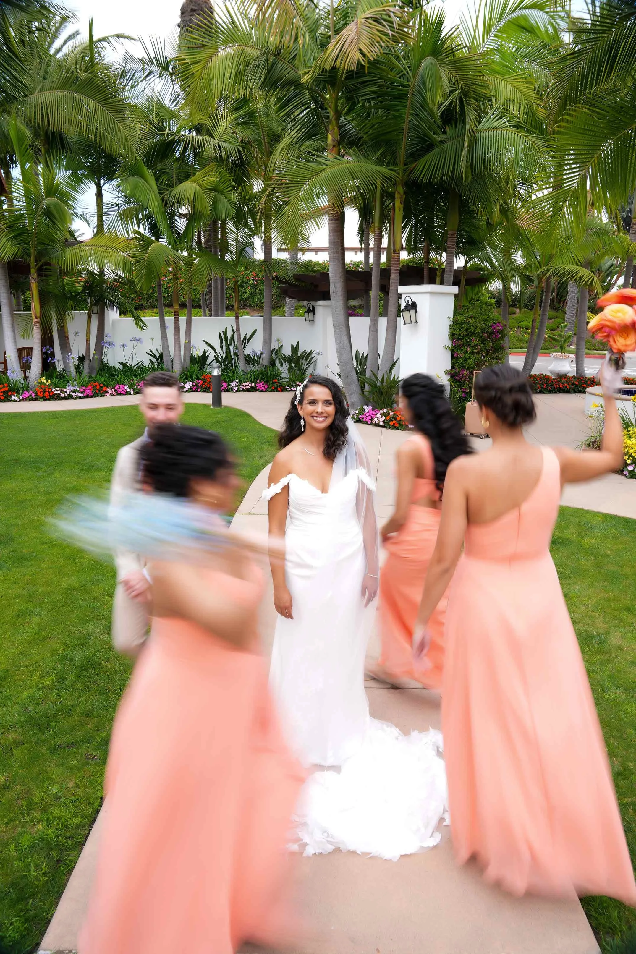 A bride in a white wedding gown stands smiling on a paved pathway surrounded by several bridesmaids wearing peach-colored dresses, with a tropical background of palm trees and white walls.


