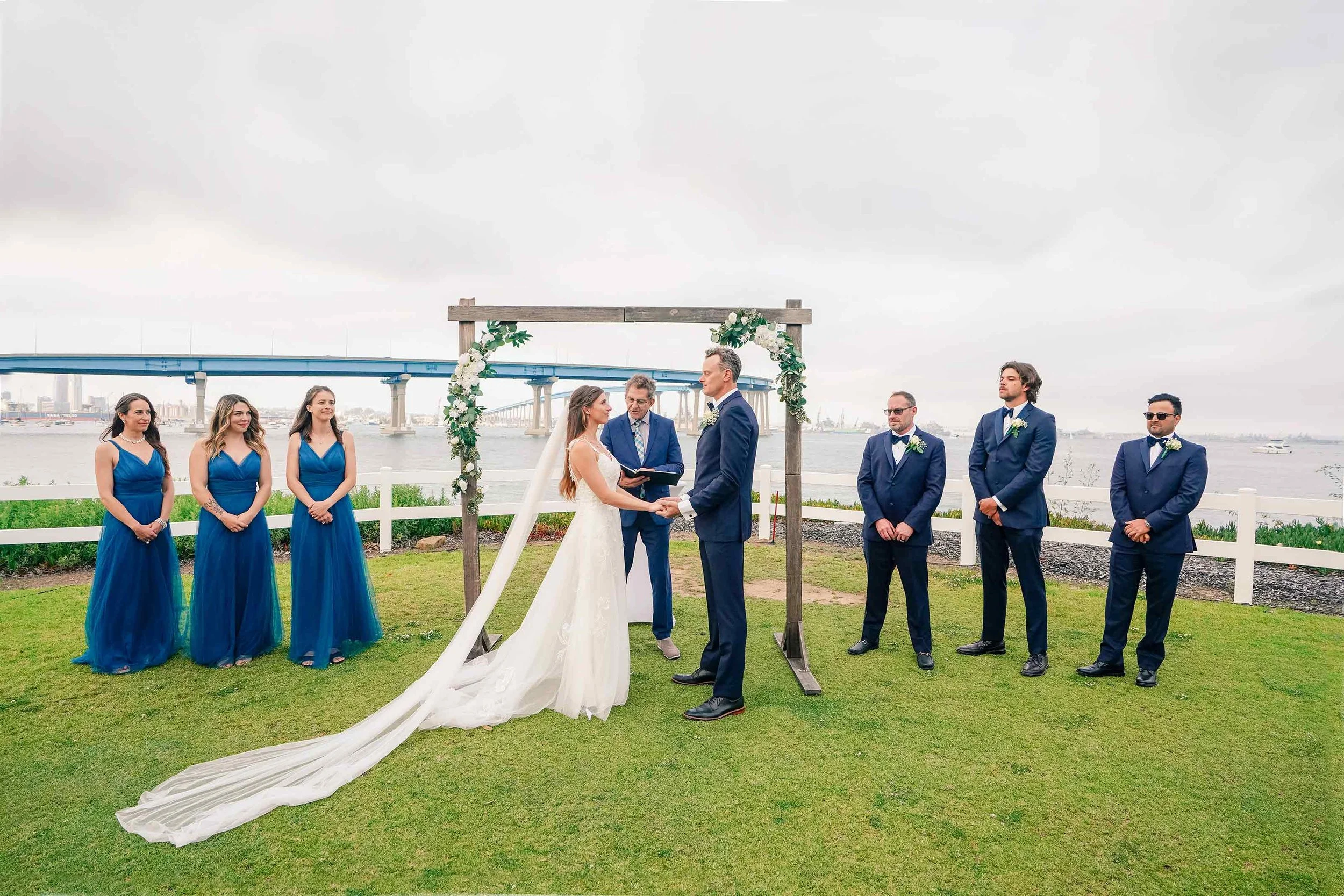 A full wedding party stands on a green lawn overlooking the water during an outdoor wedding ceremony, featuring a bride in a long gown and veil, bridesmaids in blue dresses, and groomsmen in navy blue tuxedos, with Coronado Bridge visible in the back