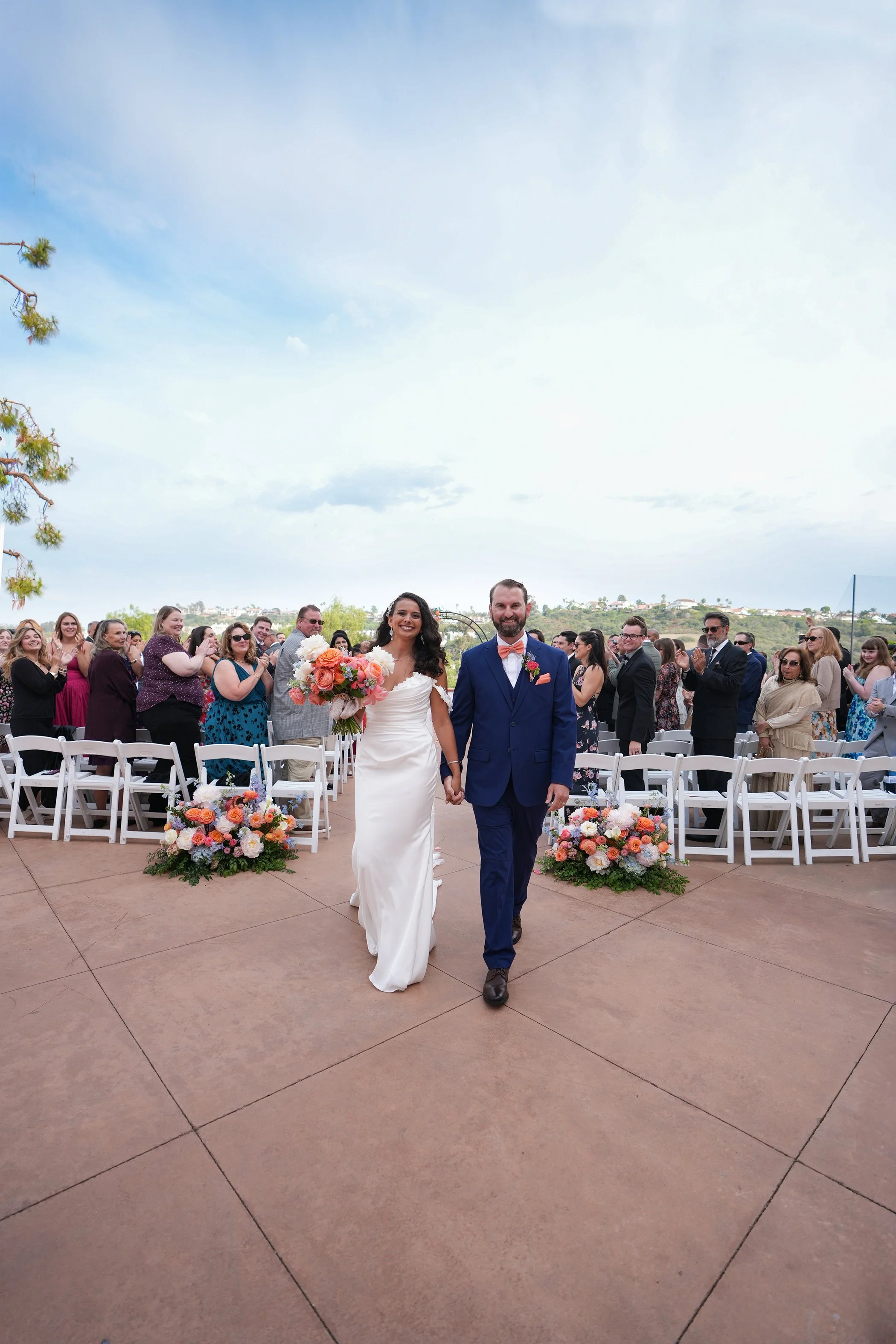 A wide-angle wedding photograph by Reimo Photography capturing a bride and groom walking hand-in-hand down the aisle after their ceremony. The couple is smiling joyfully as guests cheer from white folding chairs on a terracotta patio. Vibrant orange 