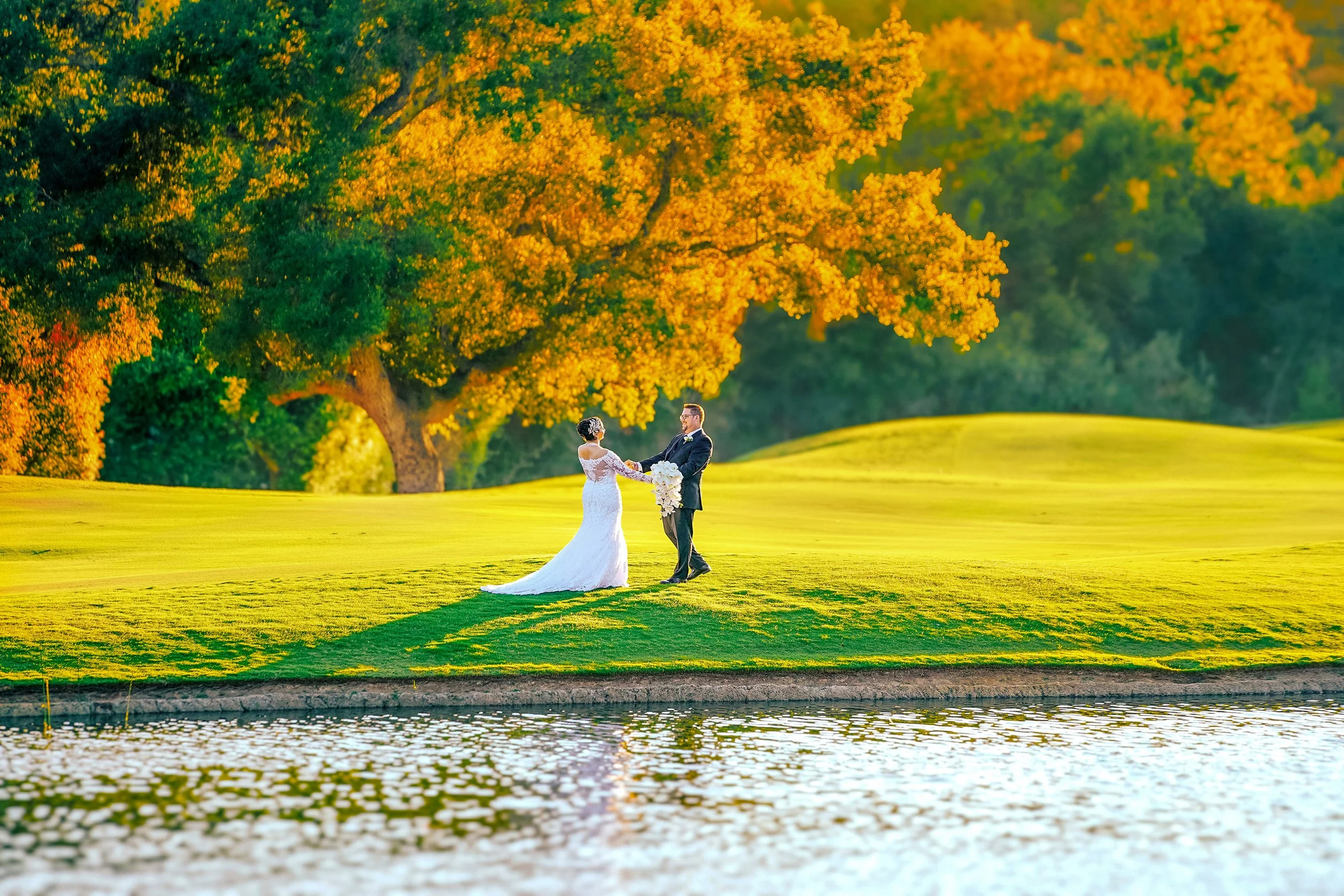 A bride and groom hold hands on a grassy hill overlooking a pond at a golf course during golden hour.