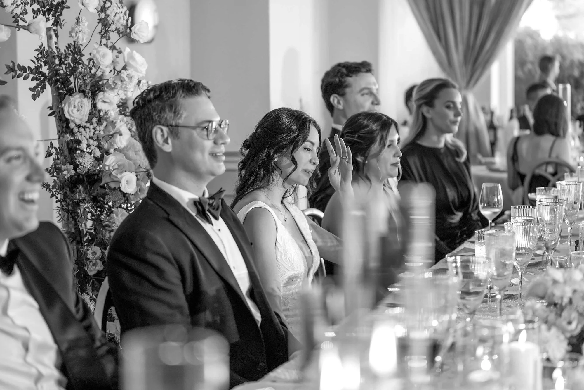 A black and white photograph of a wedding party seated at a long reception table. A man in a tuxedo smiles broadly on the left, next to a groom in a tuxedo and glasses who is also smiling. Next to him, the bride in a white dress raises her hand as sh