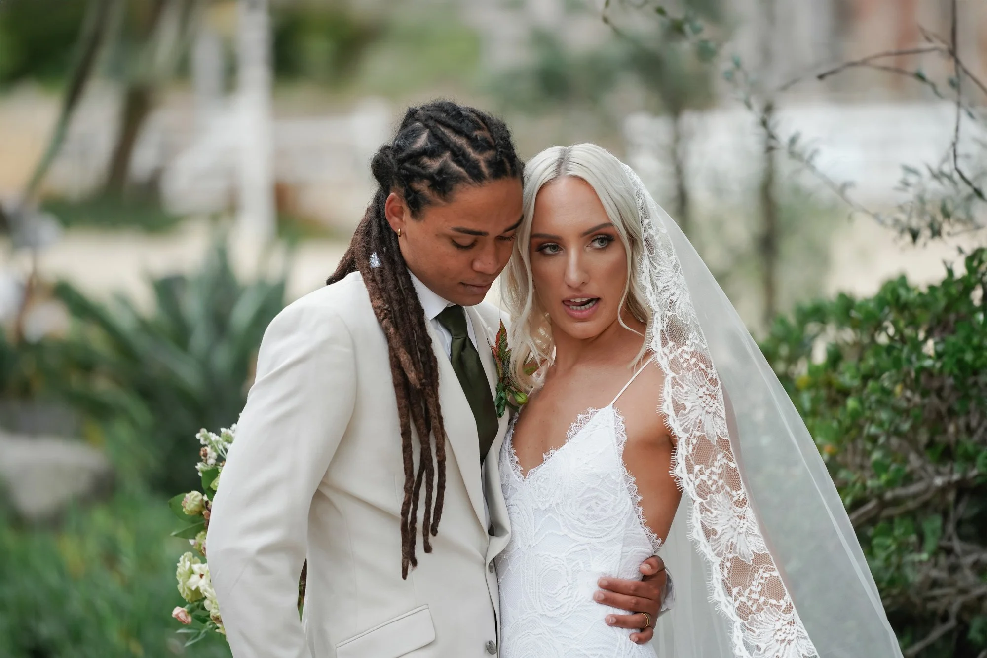 A close-up photo of the bride and groom posing together, with one wearing a cream-colored suit and olive green tie and the other in a white lace wedding dress with a long veil.


