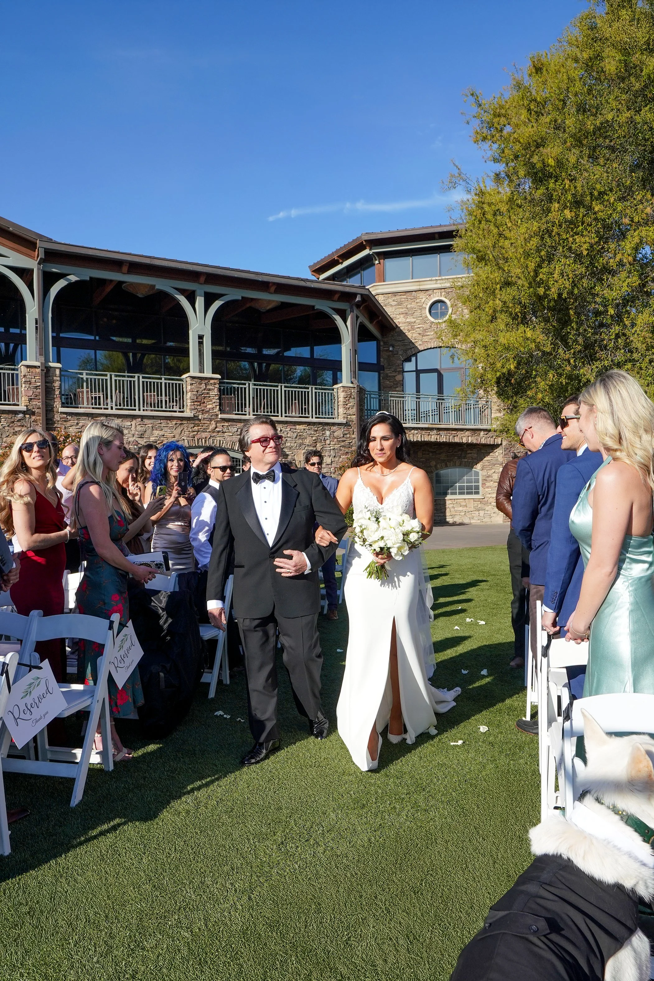 A bride and her father walk down an outdoor wedding aisle together on a bright, sunny day at Canyons Restaurant at The Crossings at Carlsbad. The bride wears a white, floor-length gown with a high leg slit and holds a large bouquet of white roses [Vi