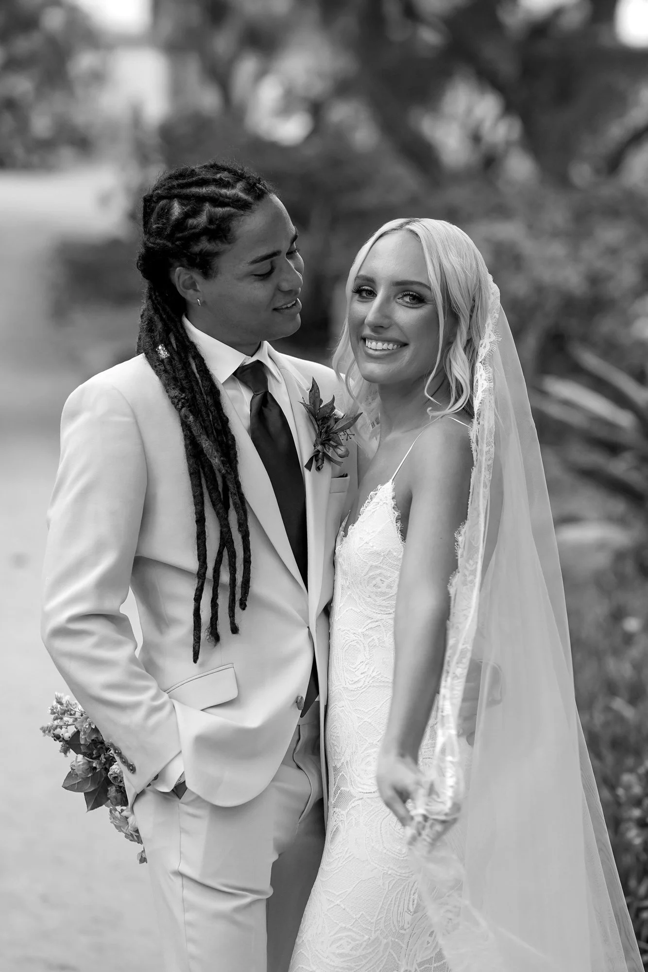 A black and white photo of a newly married couple posing for a portrait, with the groom in a cream-colored suit looking at the bride, who is in a white lace dress and veil,l and smiling at the camera.