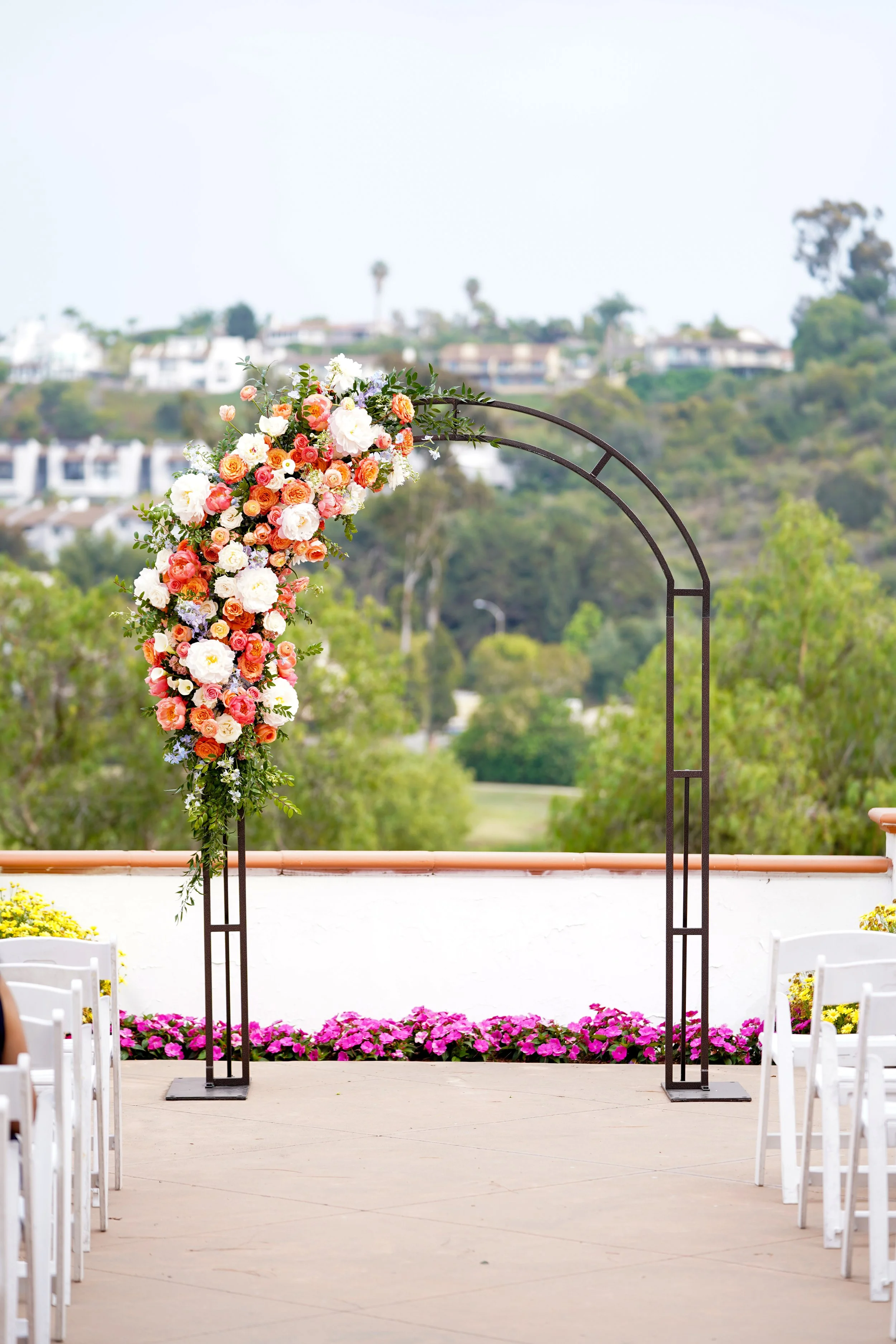 A black metal arch adorned with orange, white, and peach flowers, set up for an outdoor wedding ceremony on a patio overlooking green hills.
