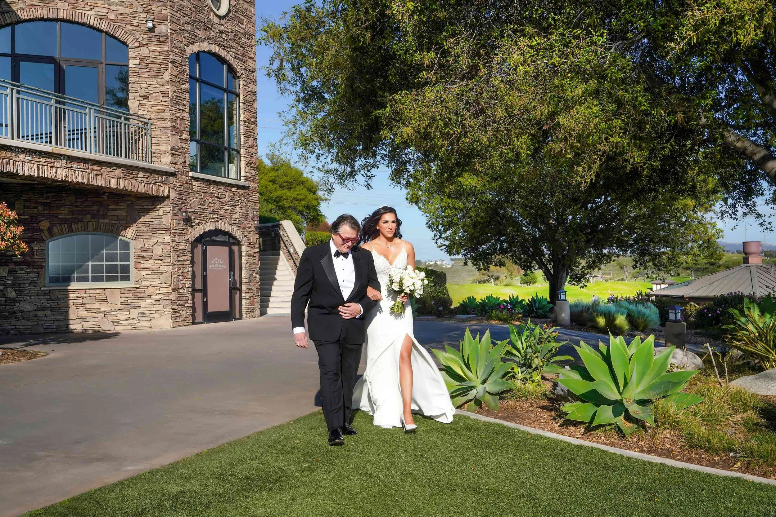 A full-length outdoor photograph of a bride in a white wedding dress with a high leg slit, holding a white floral bouquet, walking arm-in-arm with her father in a black tuxedo down a paved and grassy aisle in Carlsbad. A stone building, large green p