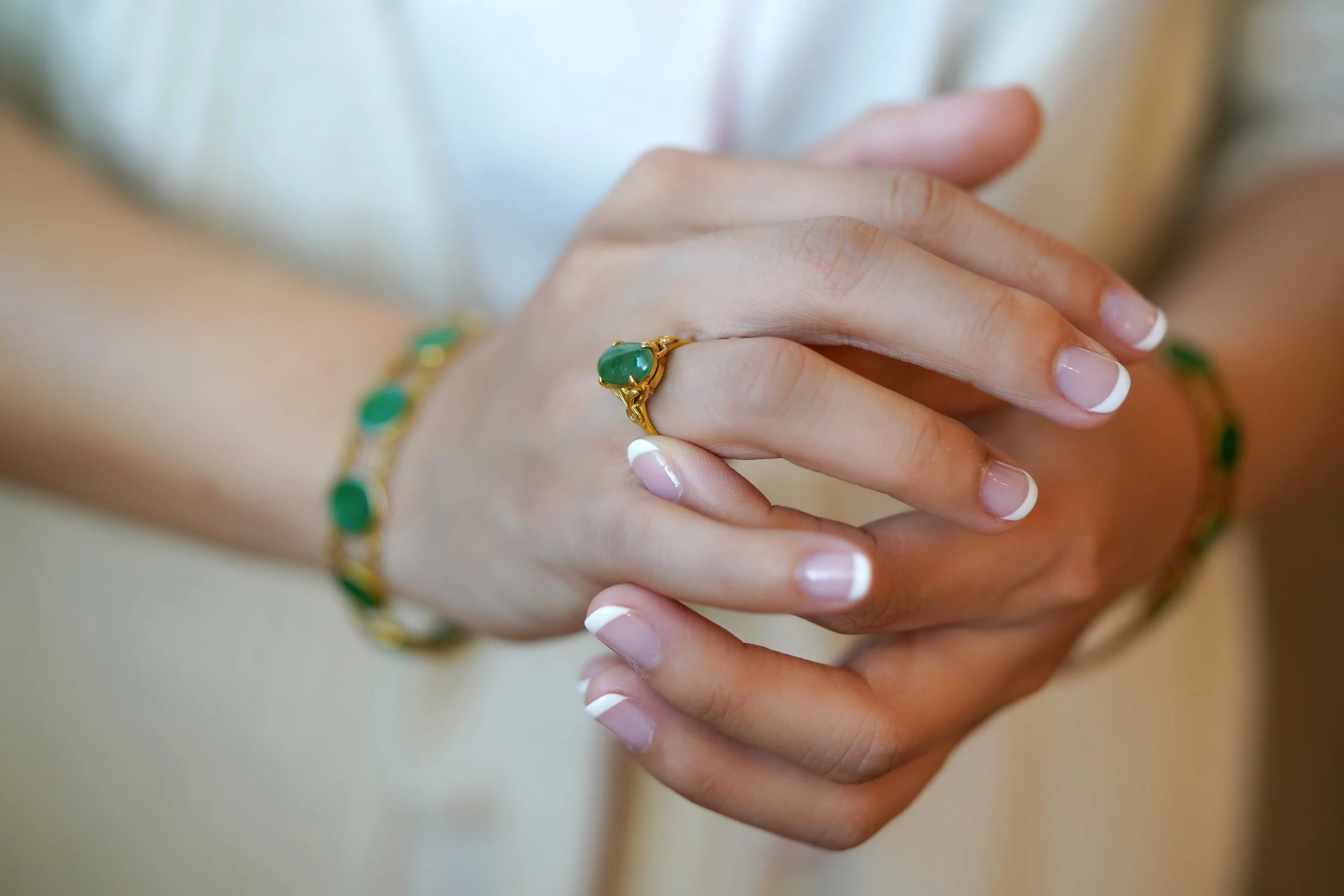 A close-up shot of a bride's hands with French manicured nails, showing a gold ring set with an oval green stone and a matching bracelet as she prepares for her wedding day.