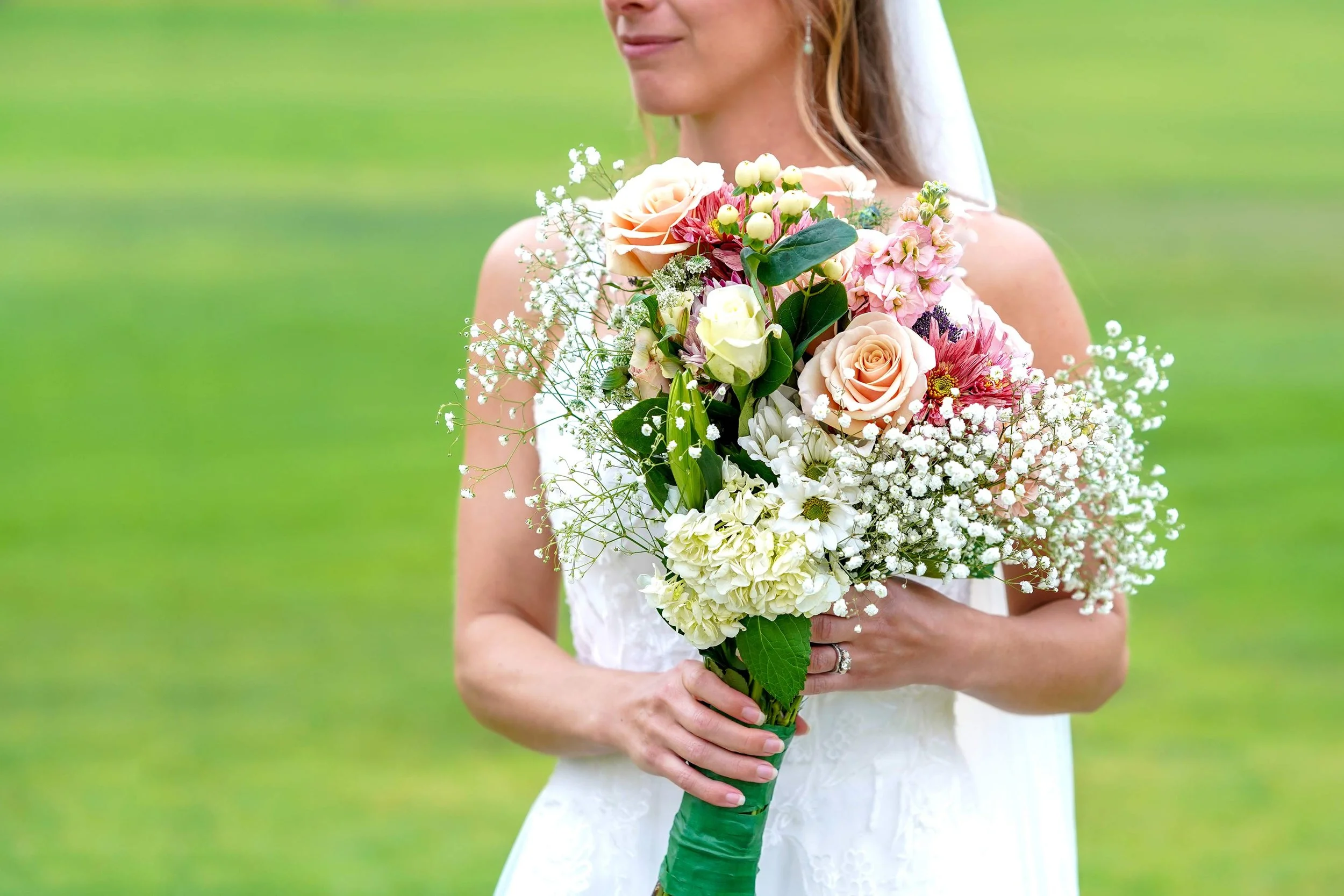 A bride in a white dress holds a vibrant bouquet of pink, peach, white, and red flowers with green foliage and baby's breath, against a bright green, blurred background.