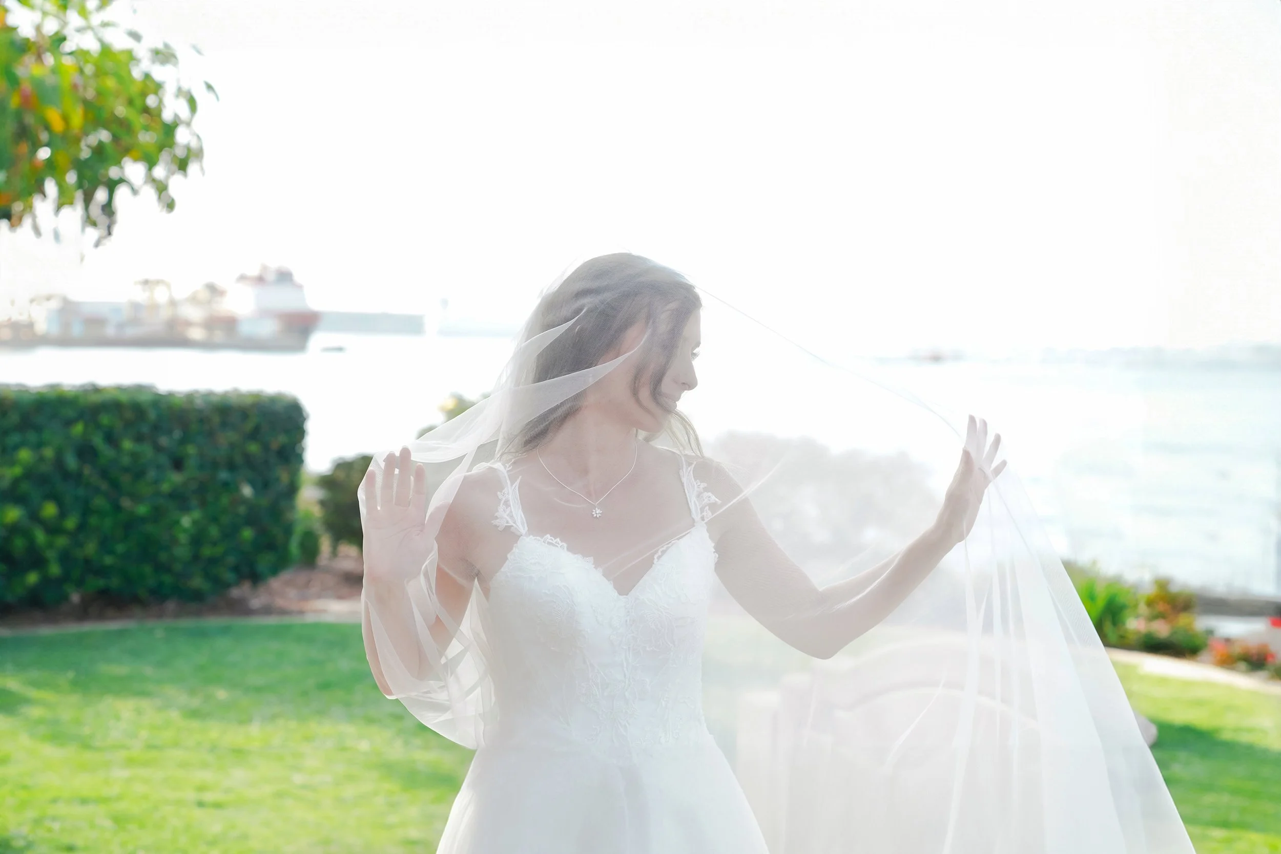 A bride in a white lace wedding gown and a long veil stands outdoors on a sunny day in Coronado, San Diego, holding the veil out as it flows in the breeze, with a body of water and a large cargo ship visible in the background.


