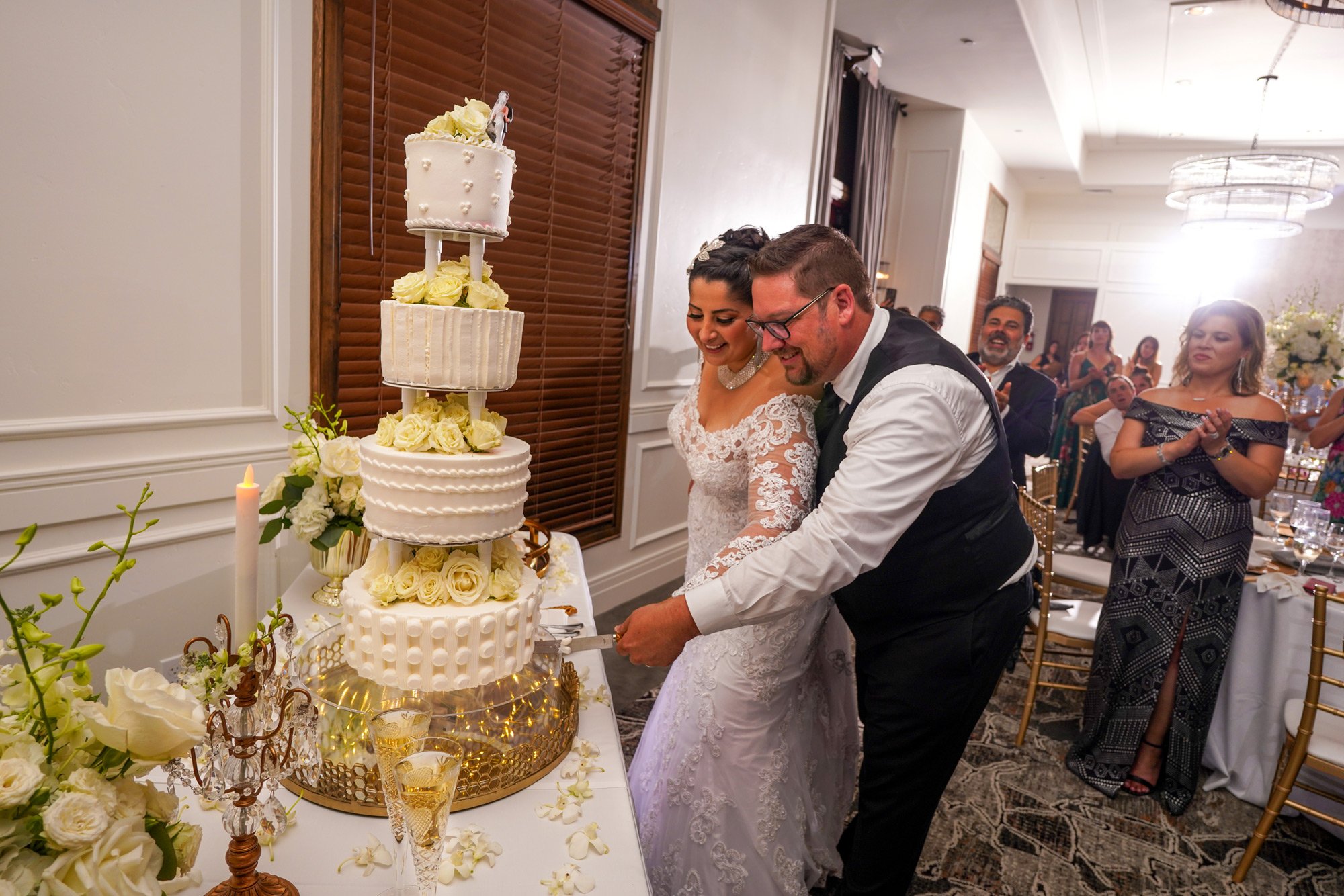 A bride in a white lace dress and a groom in a black vest and white shirt stand together, smiling as they hold a knife and cut the bottom tier of a tall, four-tiered white wedding cake adorned with yellow roses. Guests are visible watching and clappi