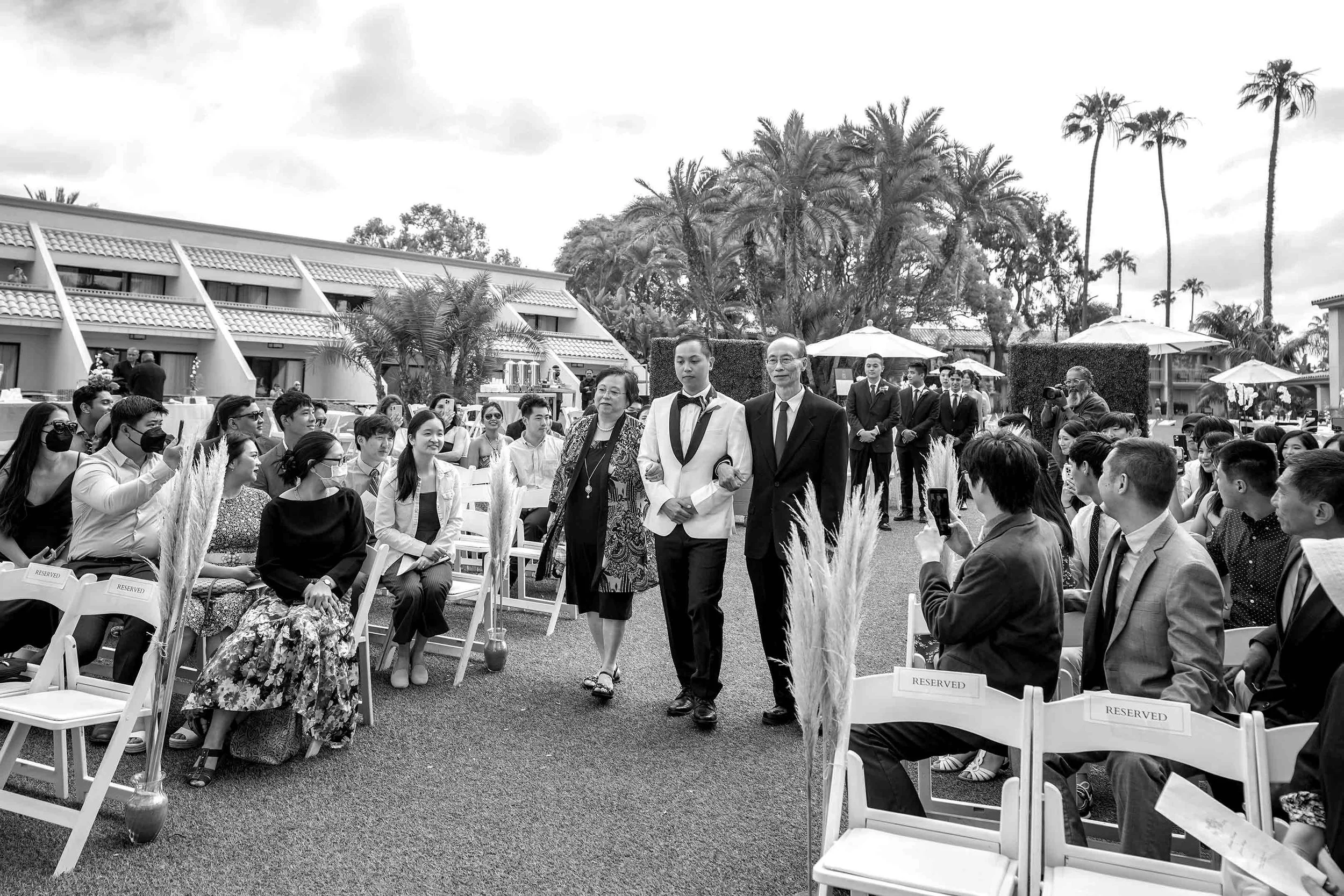 A black and white photograph of a groom in a white tuxedo jacket, escorted arm-in-arm by his mother and father, walking down an outdoor wedding aisle in Mission Bay past seated guests in white folding chairs.