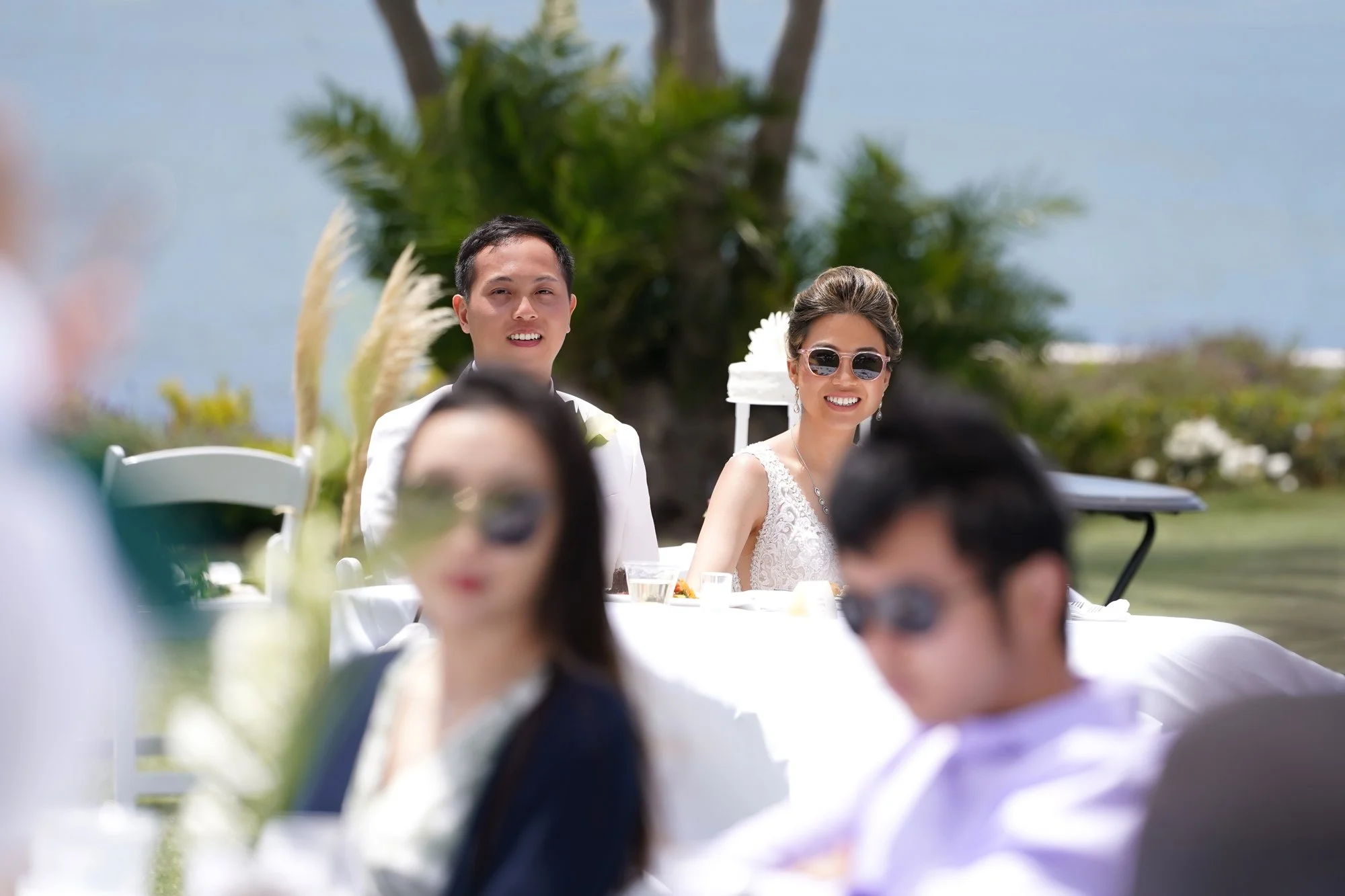 A bride in a white lace gown and sunglasses and a groom in a white tuxedo jacket sit smiling at their table during their sunny outdoor wedding reception.



