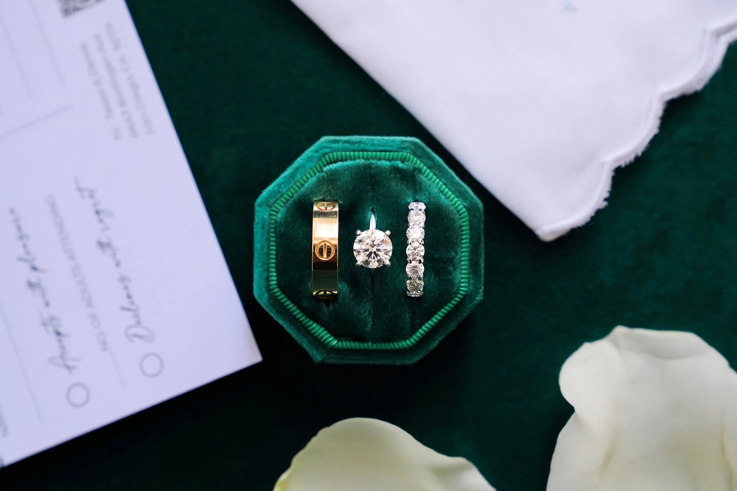A close-up photograph of three elegant rings nestled in a dark emerald-green velvet ring box. The rings include a wide gold band with screw motifs (resembling a Cartier Love ring), a round brilliant diamond solitaire engagement ring, and a diamond et