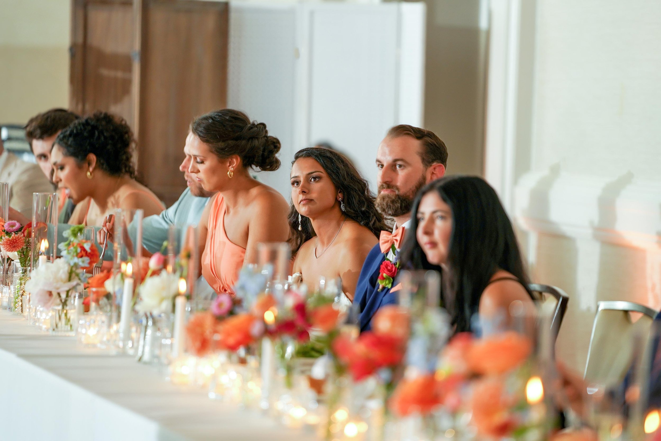 A group of well-dressed people, including a bride and groom, are seated side-by-side at a long white table adorned with colorful floral arrangements and candles, looking forward during an event like a wedding reception.