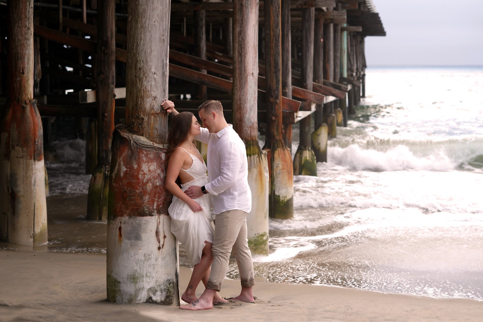 An outdoor photograph of a couple embracing and about to kiss on a sandy beach in Pacific Beach, standing under a large wooden pier as waves crash nearby. The woman wears a white sundress, and the man wears a white shirt and tan pants; both are baref