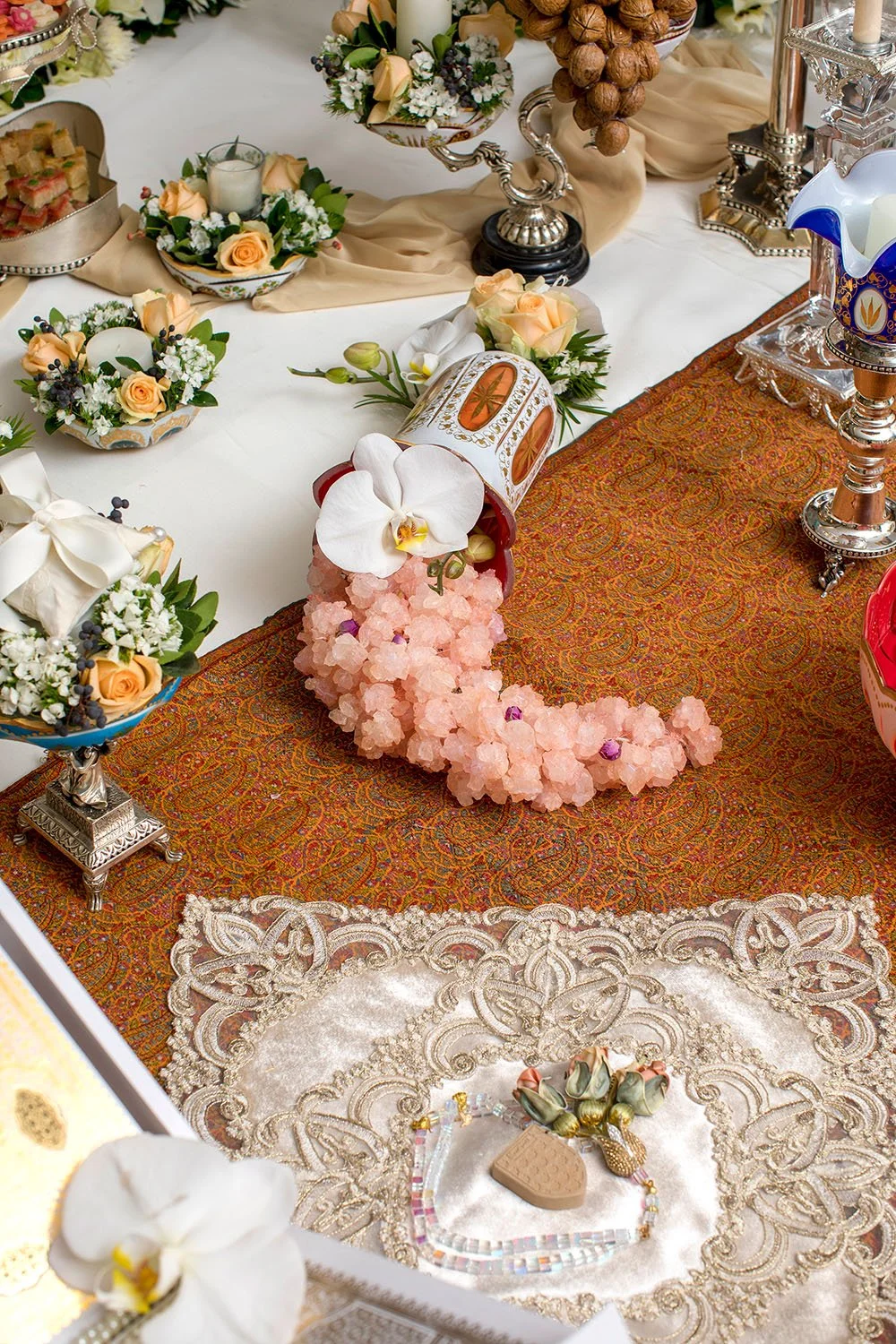 A close-up, overhead view of a traditional Persian wedding table (Sofreh Aghd), highlighting a display of pink rock candy flowing from a white and orange ceramic vase, with white orchids, silver serving dishes, fresh flowers, and an orange patterned 